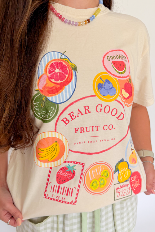 A person wearing a cream-colored graphic t-shirt with a colorful fruit-themed print that reads 'BEAR GOOD FRUIT CO.' and 'FAVOR THAT REMAINS.