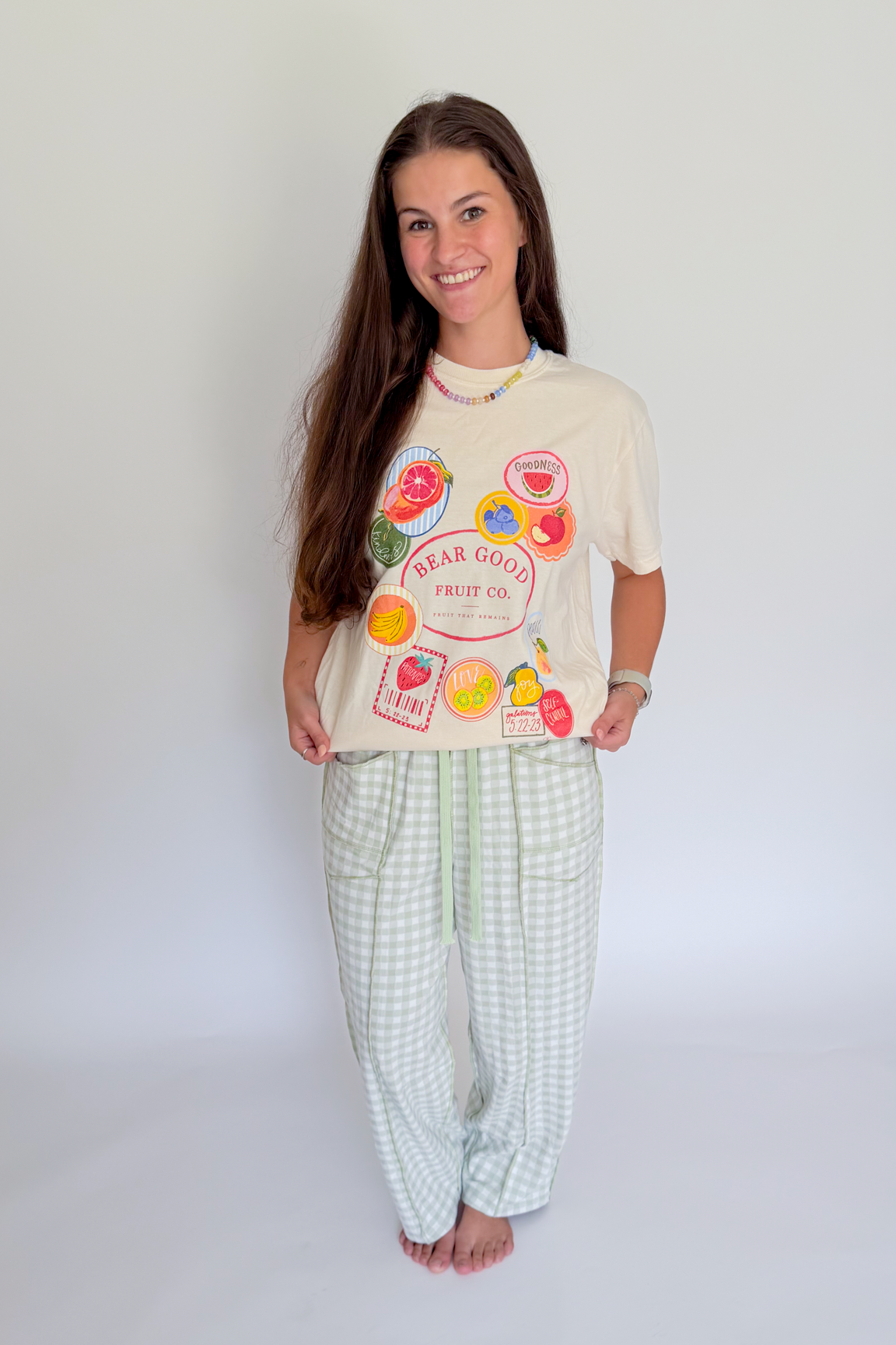 A woman modeling a two-piece pajama set consisting of a cream-colored graphic t-shirt with fruit illustrations and light green and white checkered pants, standing against a plain white studio background.