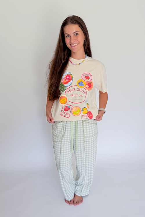 A woman modeling a two-piece pajama set consisting of a cream-colored graphic t-shirt with fruit illustrations and light green and white checkered pants, standing against a plain white studio background.