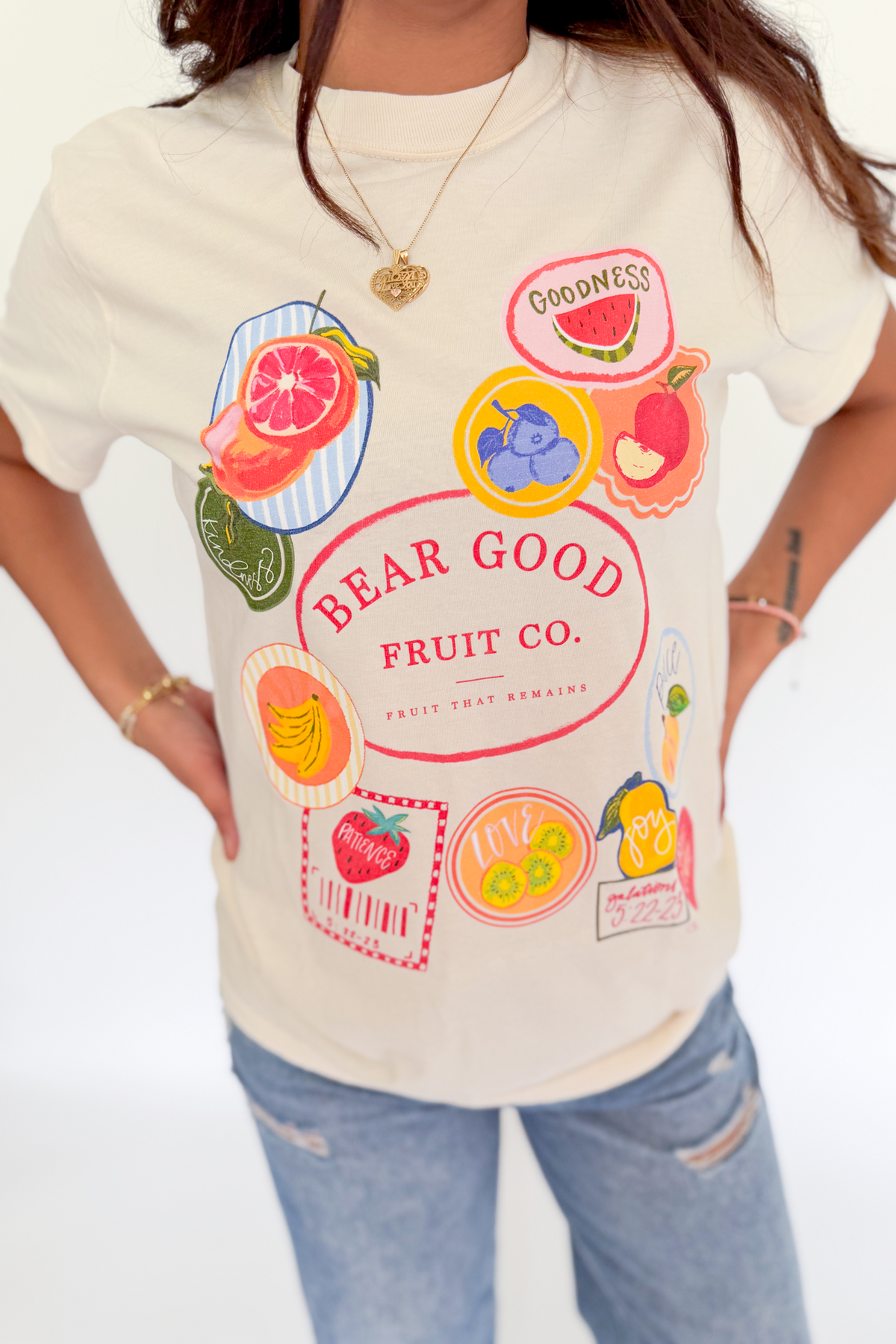 A woman wearing a cream-colored graphic t-shirt with colorful fruit illustrations and the text 'BEAR GOOD FRUIT CO.', paired with light blue jeans.