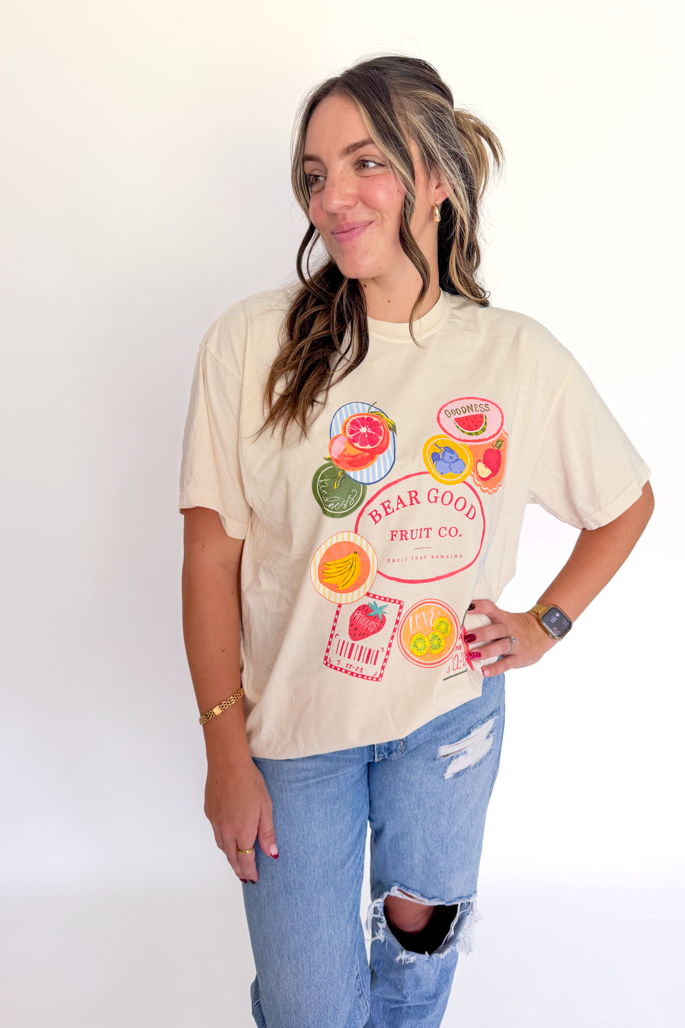 A woman modeling a beige graphic t-shirt with a colorful fruit-themed print that reads 'BEAR GOOD FRUIT CO.' and 'SWEET', paired with light blue ripped jeans, against a plain white background.