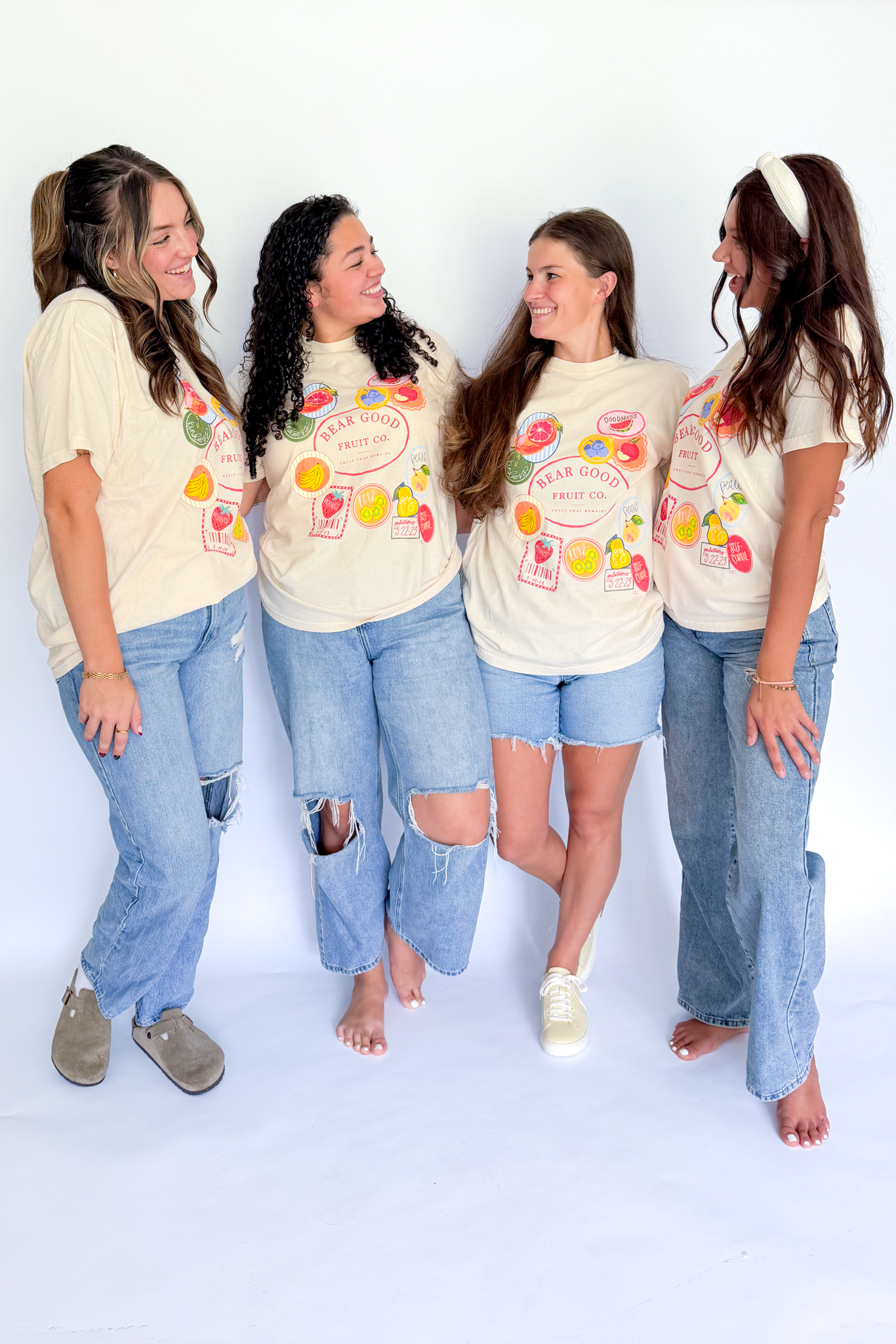 Four women modeling matching cream graphic t-shirts with colorful fruit illustrations and the text 'Fruit of the Loom' paired with light-wash denim bottoms, standing against a plain white studio background.