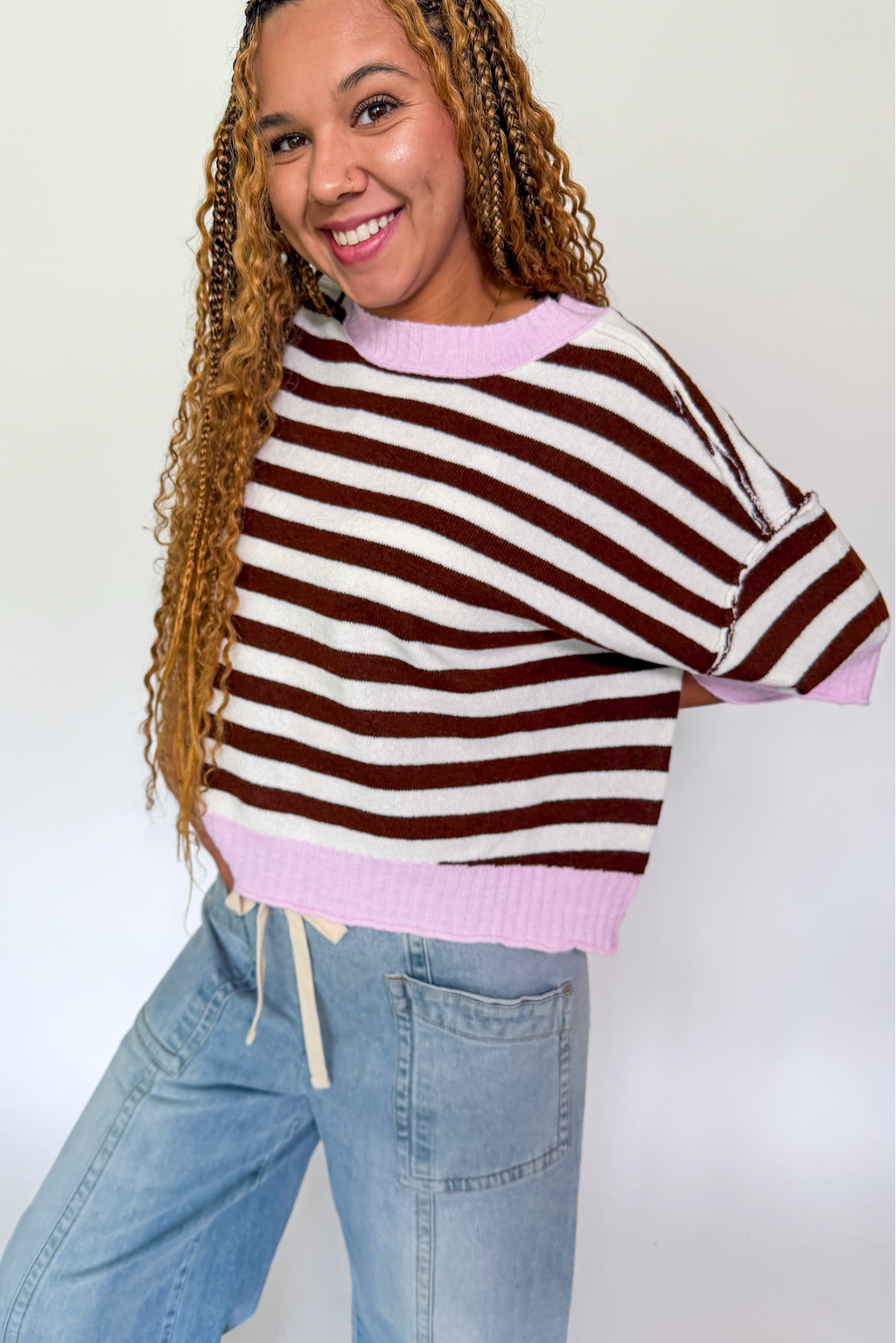A woman wearing a brown and white striped knit sweater with light pink ribbed trim, paired with light blue jeans, posing against a plain white background.