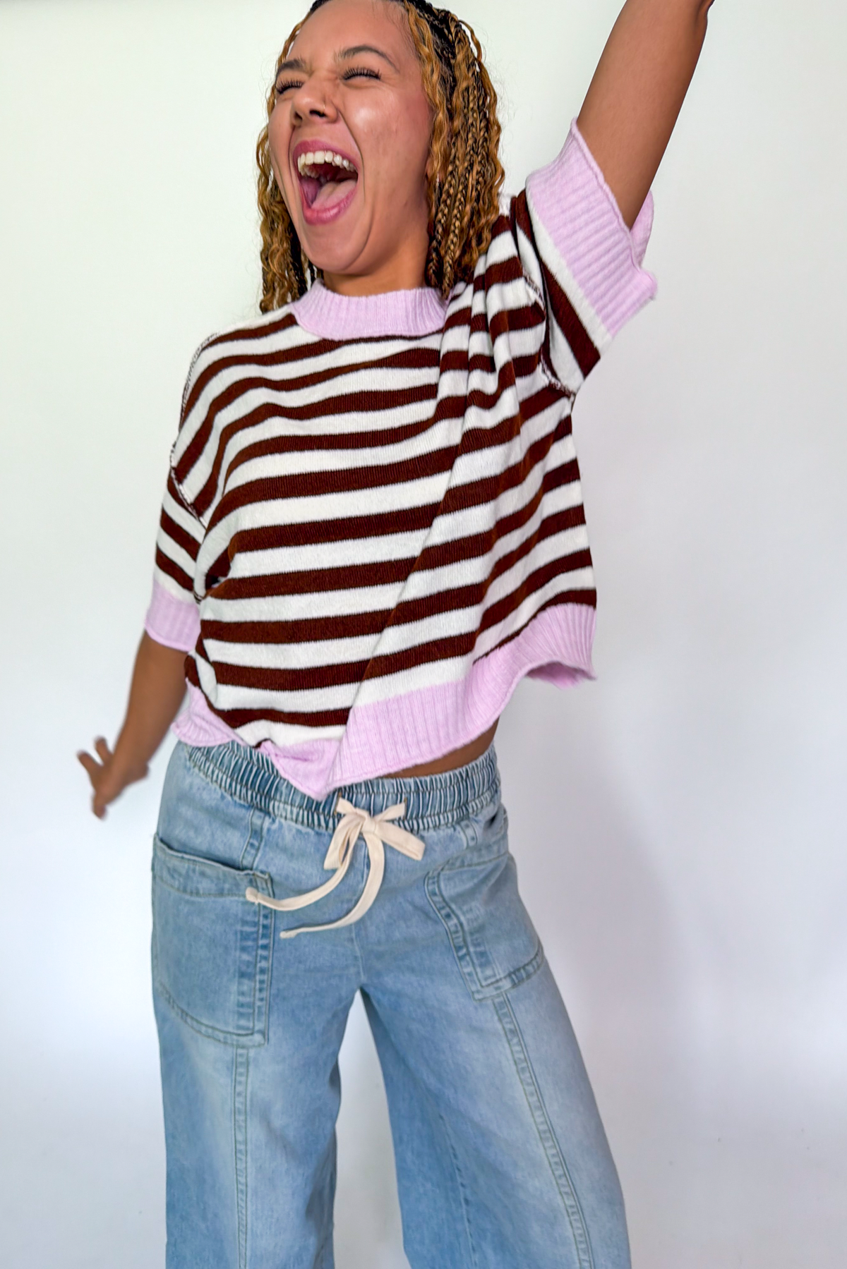 A woman wearing a brown and white striped knit sweater with light pink ribbed trim and light blue jeans, posing with one arm raised against a plain white background.