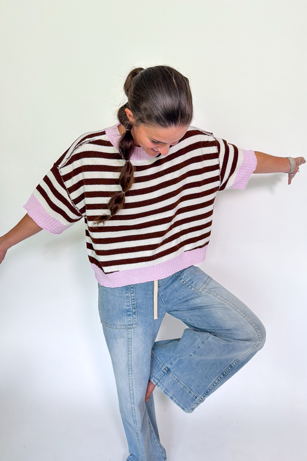 A woman wearing a brown and white striped short-sleeve knit sweater with light pink ribbed trim, paired with light blue jeans, posing against a plain white wall.