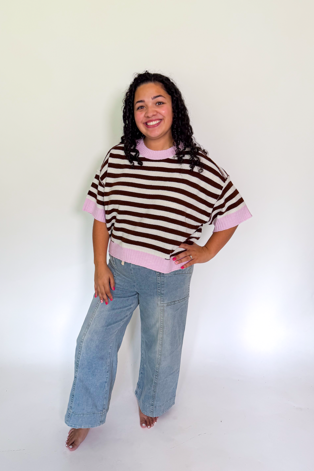 A woman modeling a brown and pink striped short-sleeve knit top with light blue wide-leg jeans, standing against a plain white studio background.