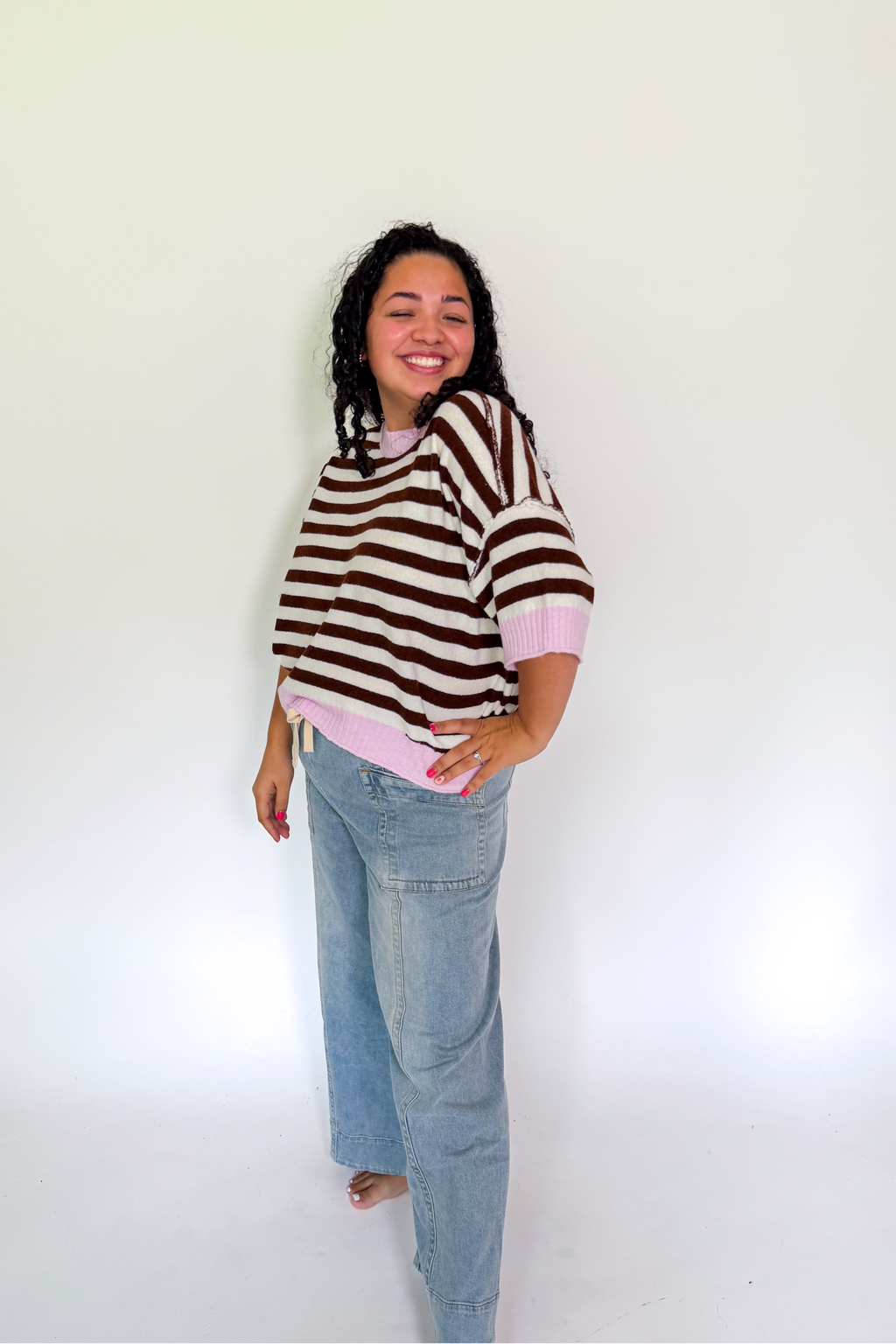 A woman modeling a brown and white striped short-sleeve top with light pink trim and light-wash blue jeans, standing against a plain white studio background.