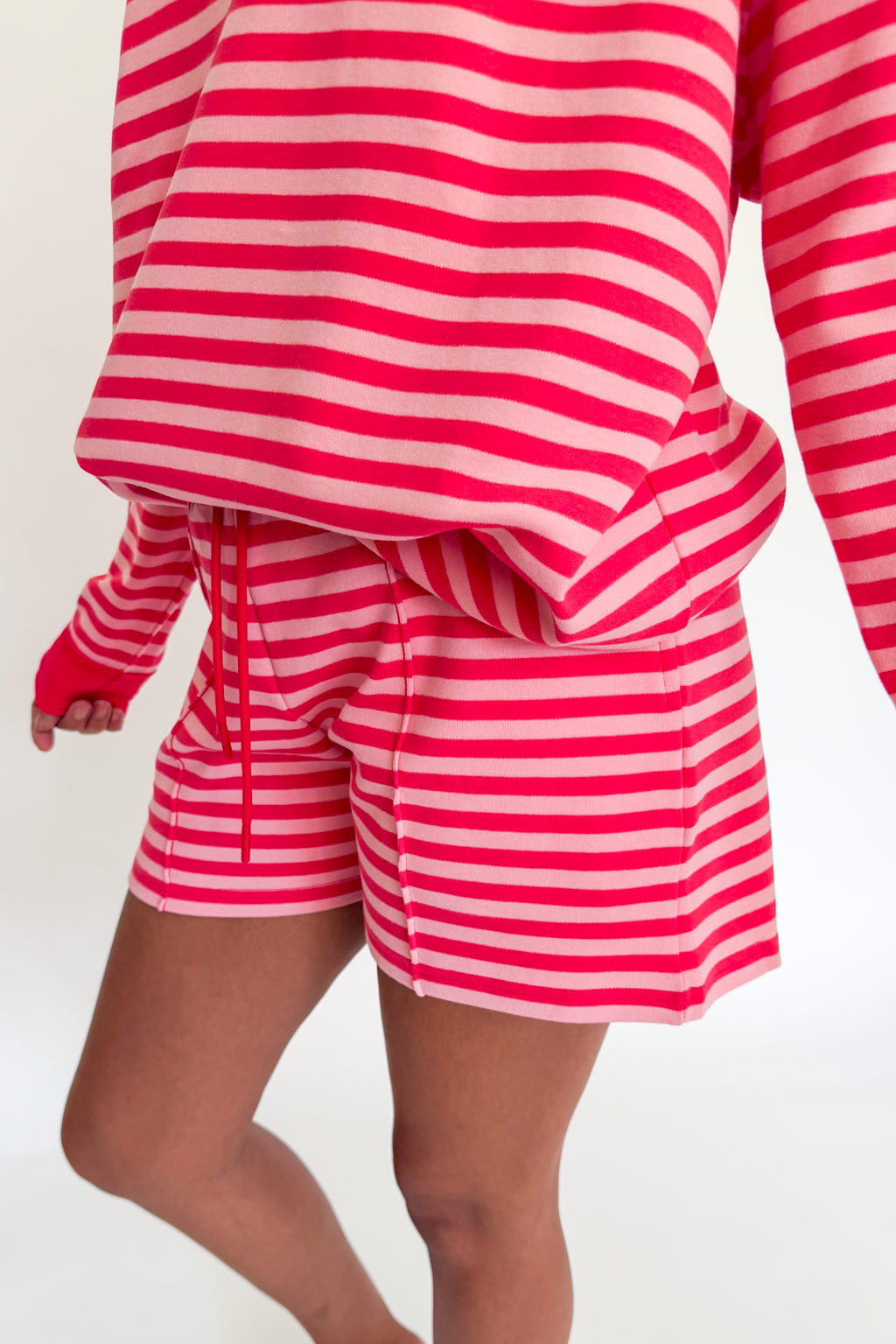 A person wearing a matching pink and white striped lounge set, featuring a cropped long-sleeve top and drawstring shorts, shown against a plain white background.