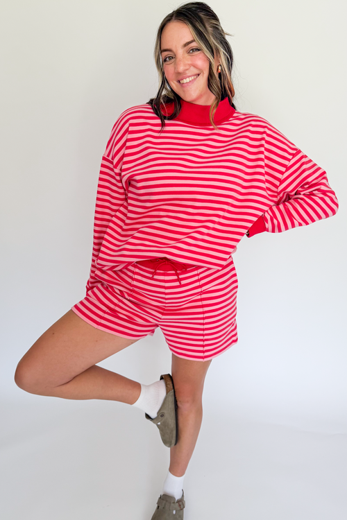 A woman modeling a matching red and white striped lounge set, featuring a long-sleeve mock neck top and shorts, paired with white socks and brown slip-on shoes, against a plain white studio background.