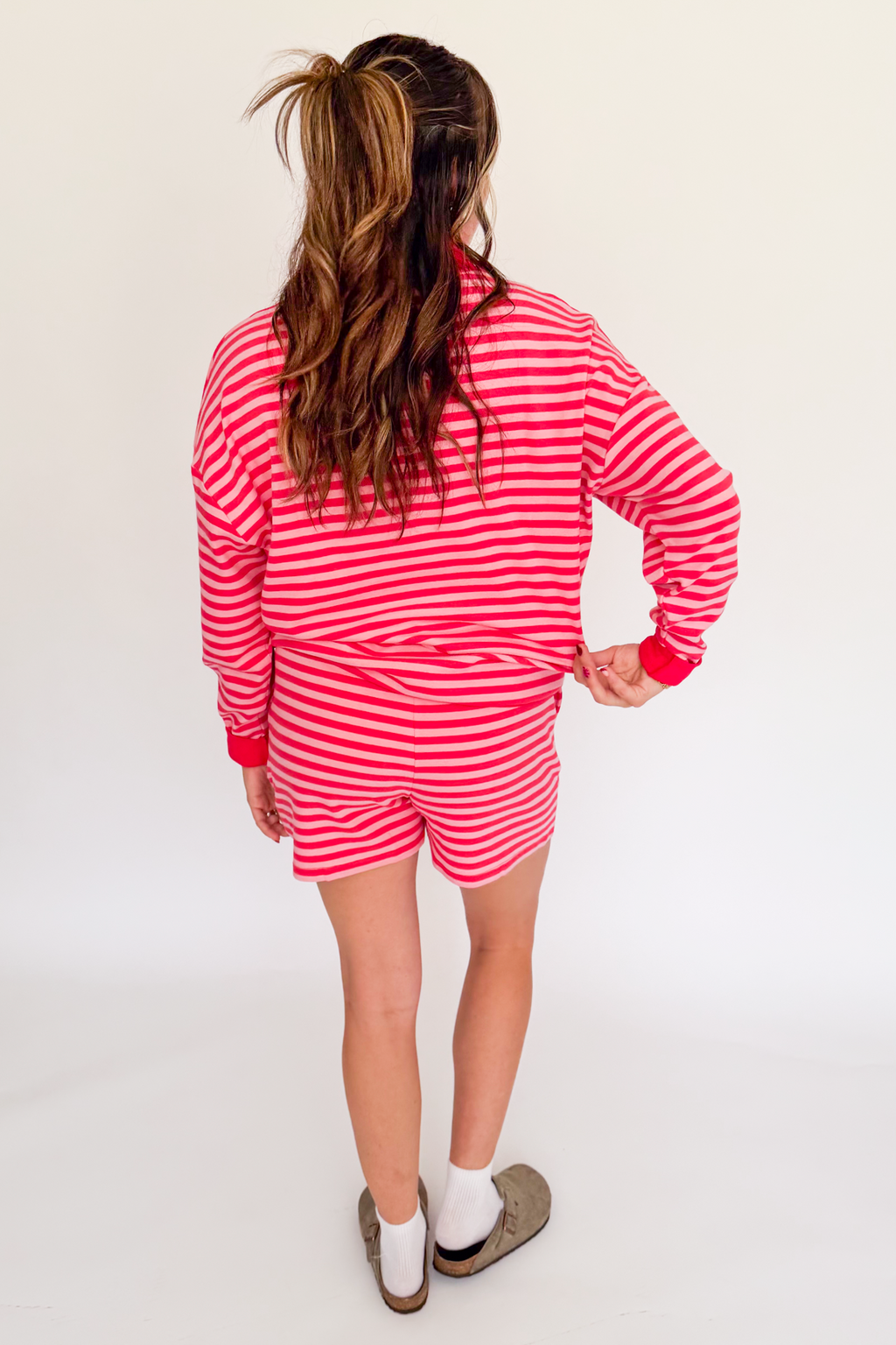 A woman viewed from behind wearing a red and white striped long-sleeve top and matching striped shorts, standing against a plain white studio background.