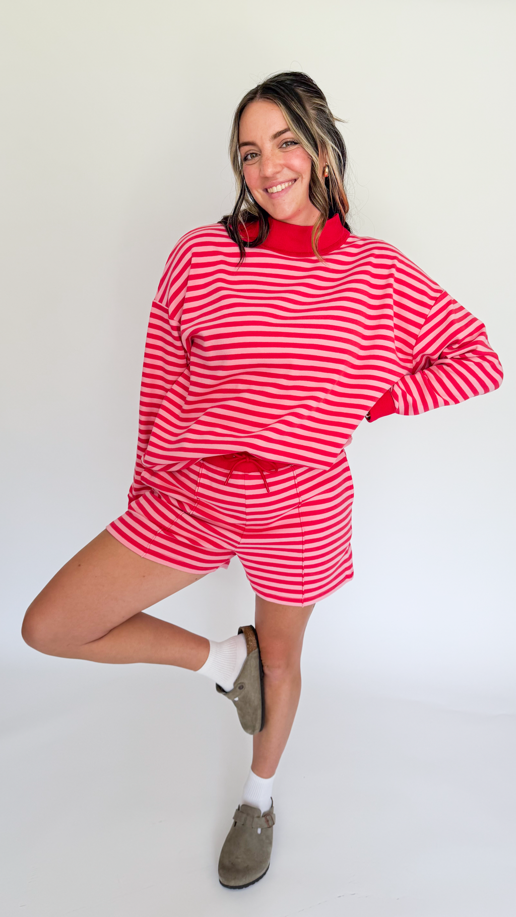 A woman modeling a matching red and white striped lounge set, featuring a long-sleeve mock neck top and shorts, paired with white socks and brown slip-on shoes, against a plain white studio background.