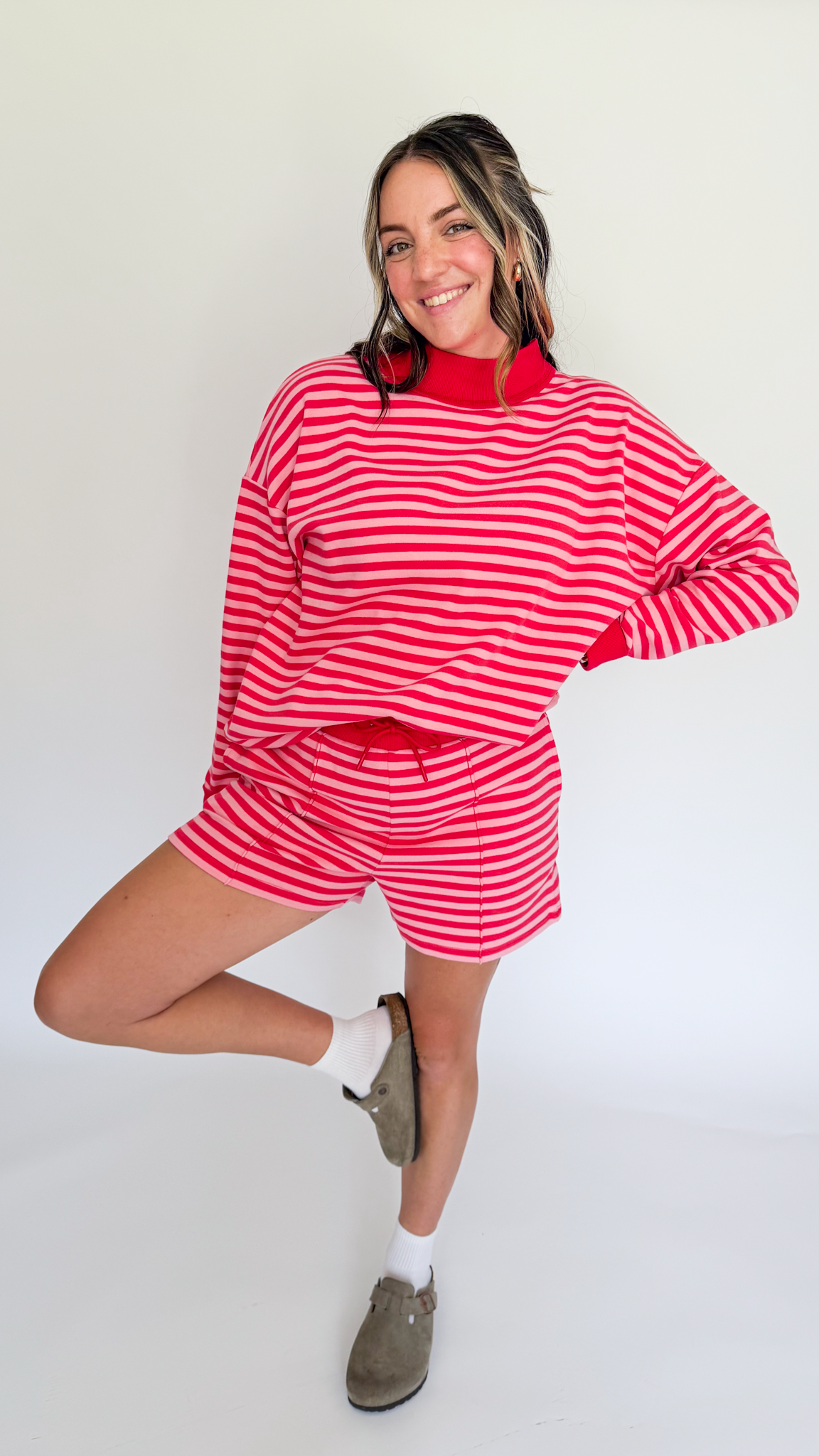 A woman modeling a matching red and white striped lounge set, featuring a long-sleeve mock neck top and shorts, paired with white socks and brown slip-on shoes, against a plain white studio background.