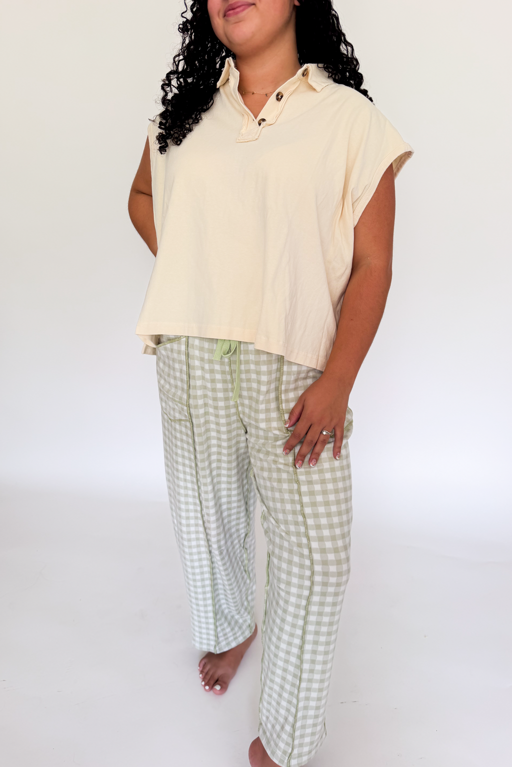 A woman modeling a light cream short-sleeve collared top and light green and white gingham pajama pants against a plain white studio background.