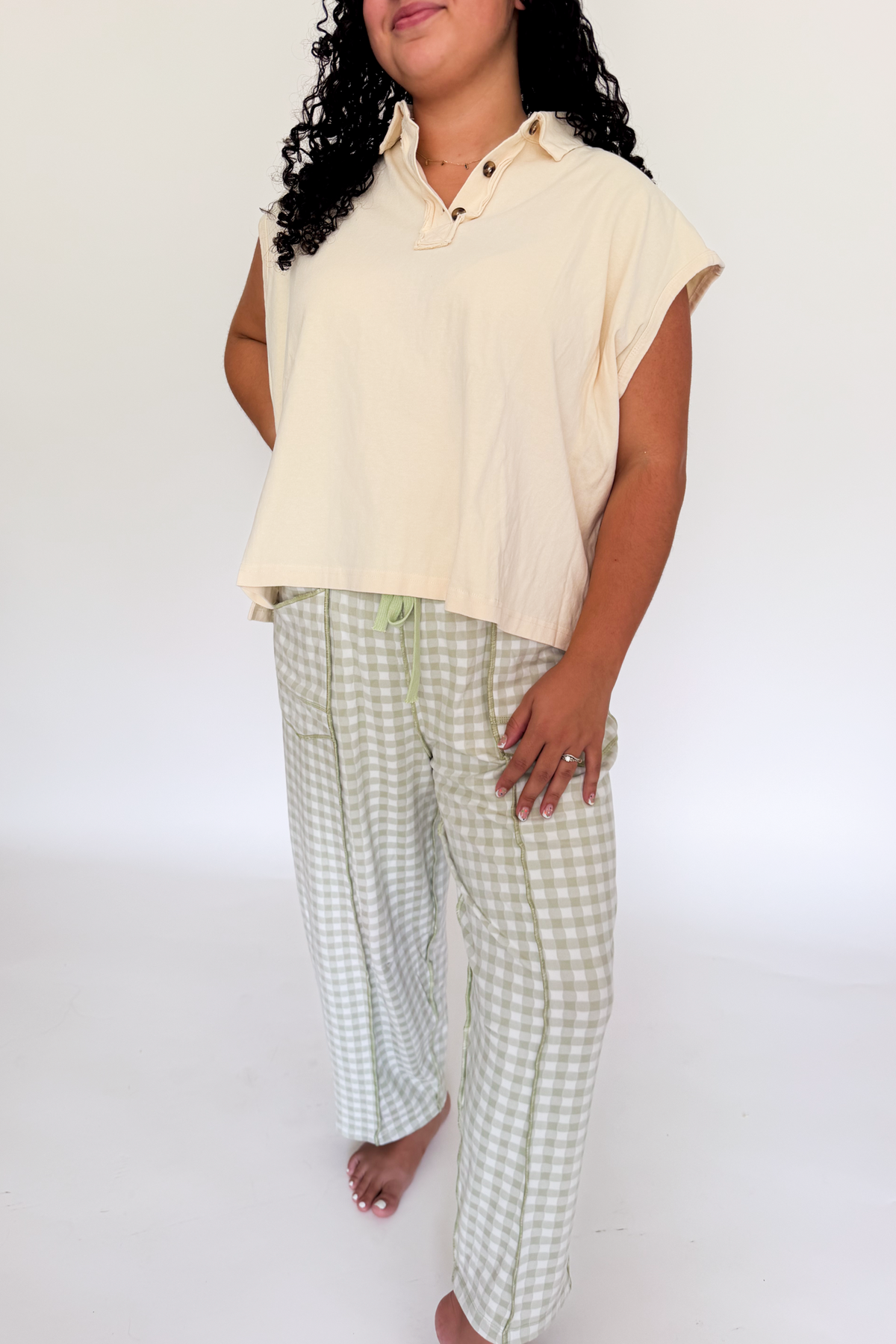 A woman modeling a light cream short-sleeve collared top and light green and white gingham pajama pants against a plain white studio background.