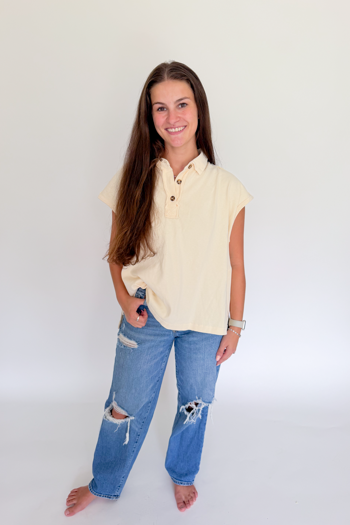 A young woman with long brown hair stands barefoot against a plain white studio background, wearing a pale yellow short-sleeve button-up shirt and distressed blue jeans.