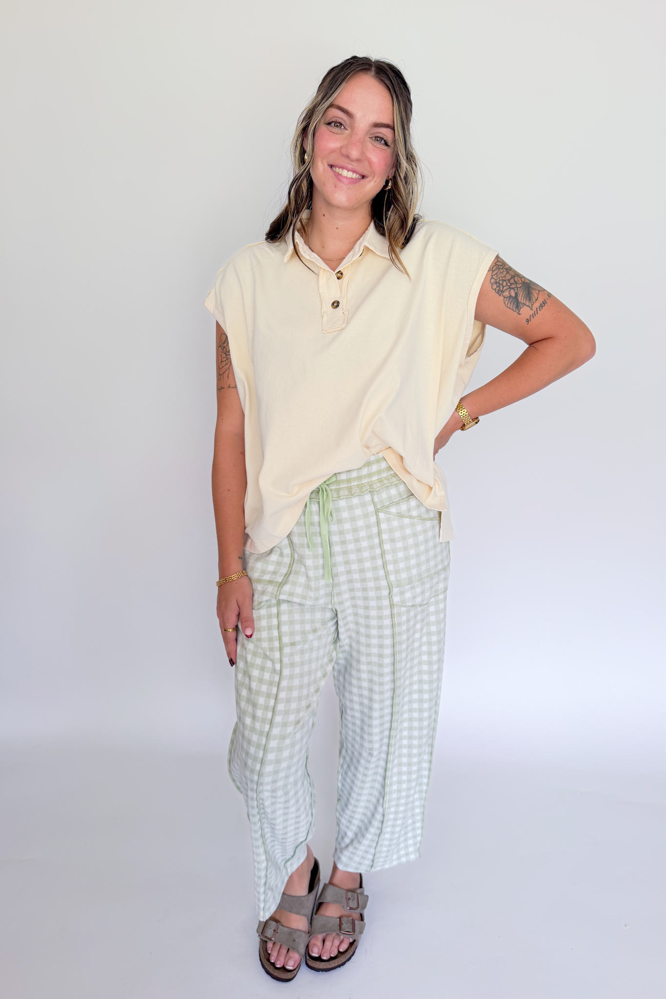 A woman modeling a light yellow short-sleeve polo shirt and light green and white gingham wide-leg pants, paired with brown sandals, against a plain white studio background.
