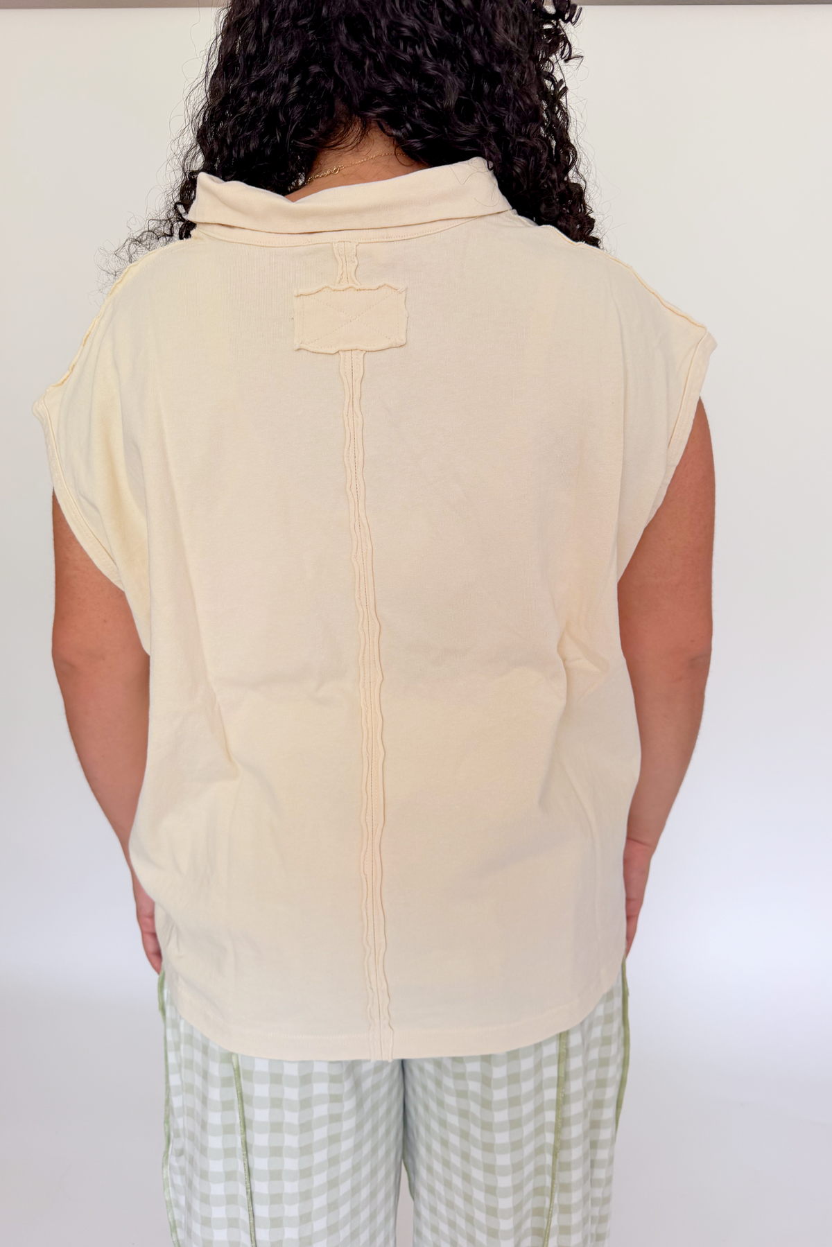 A person shown from the back wearing a cream-colored sleeveless collared top and green-and-white gingham pants against a plain white studio background.