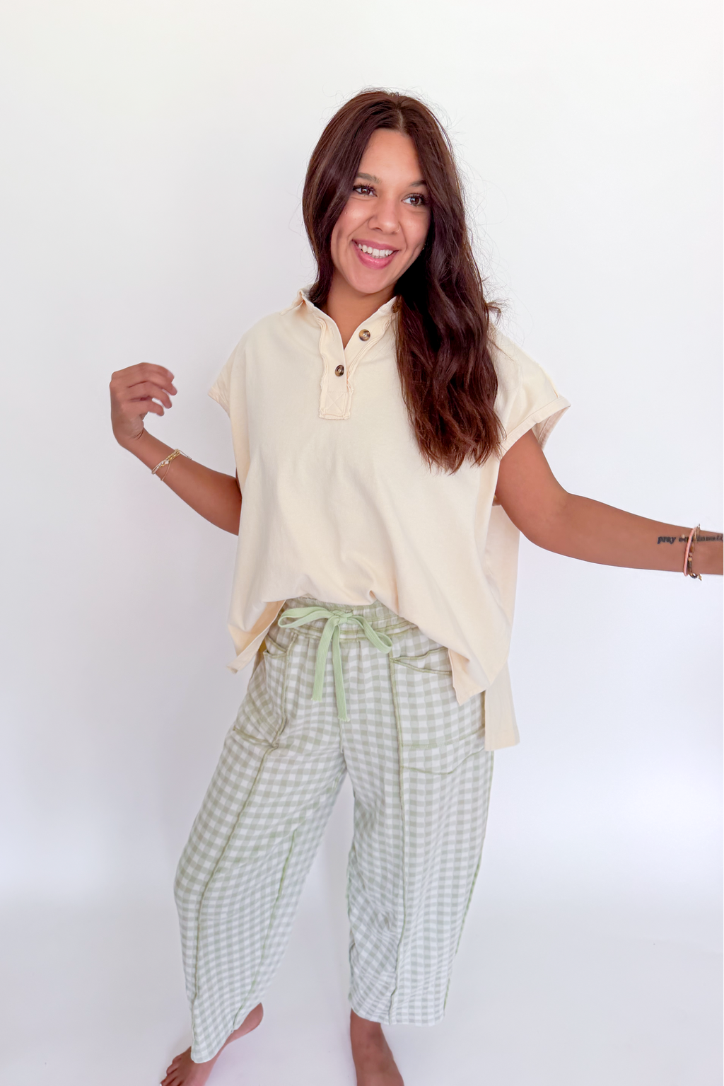 A woman posing in a light cream short-sleeve henley shirt and green and white gingham cropped pants against a plain white background.
