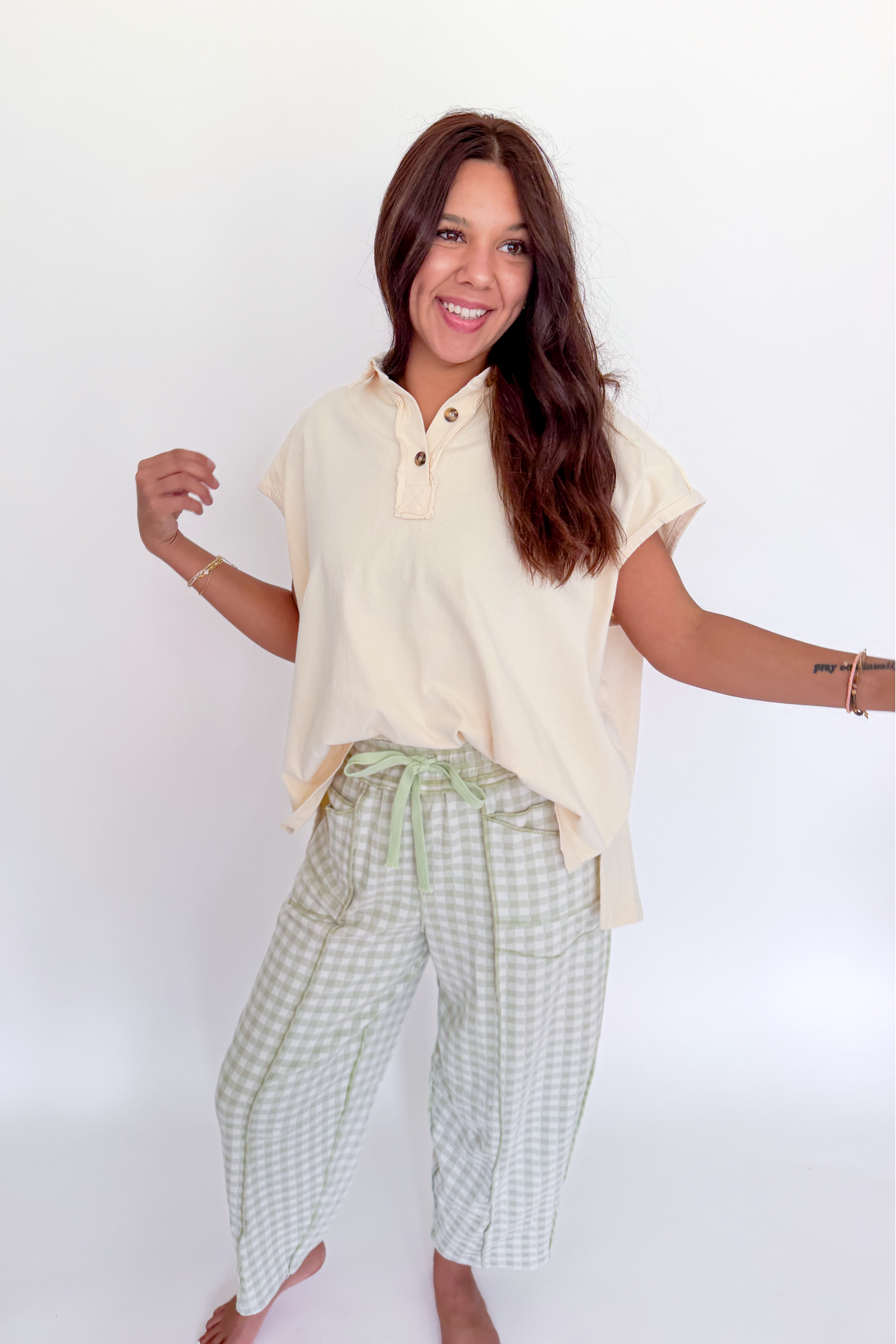 A woman posing in a light cream short-sleeve henley shirt and green and white gingham cropped pants against a plain white background.