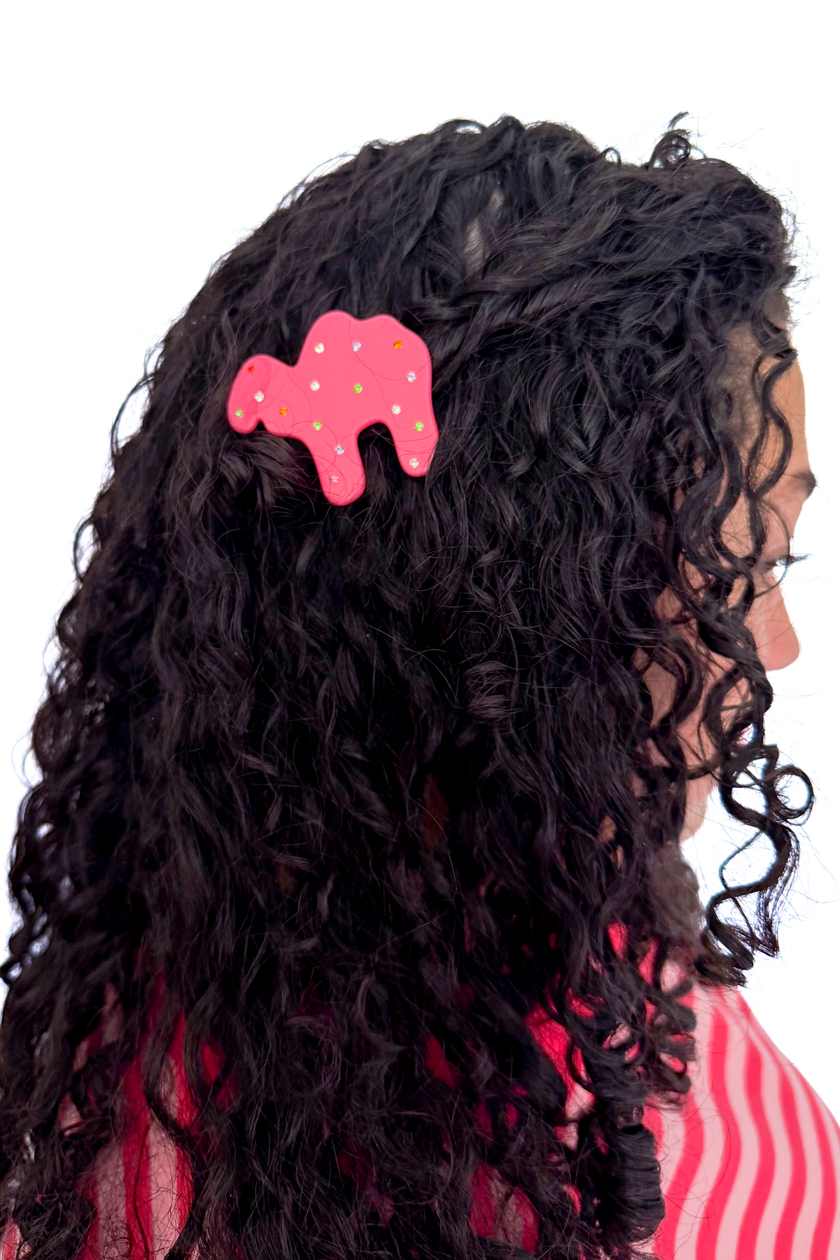 A close-up side view of a person with long, curly black hair wearing a pink camel-shaped hair clip with white polka dots. The person is also wearing a pink and white striped top, set against a plain white background.