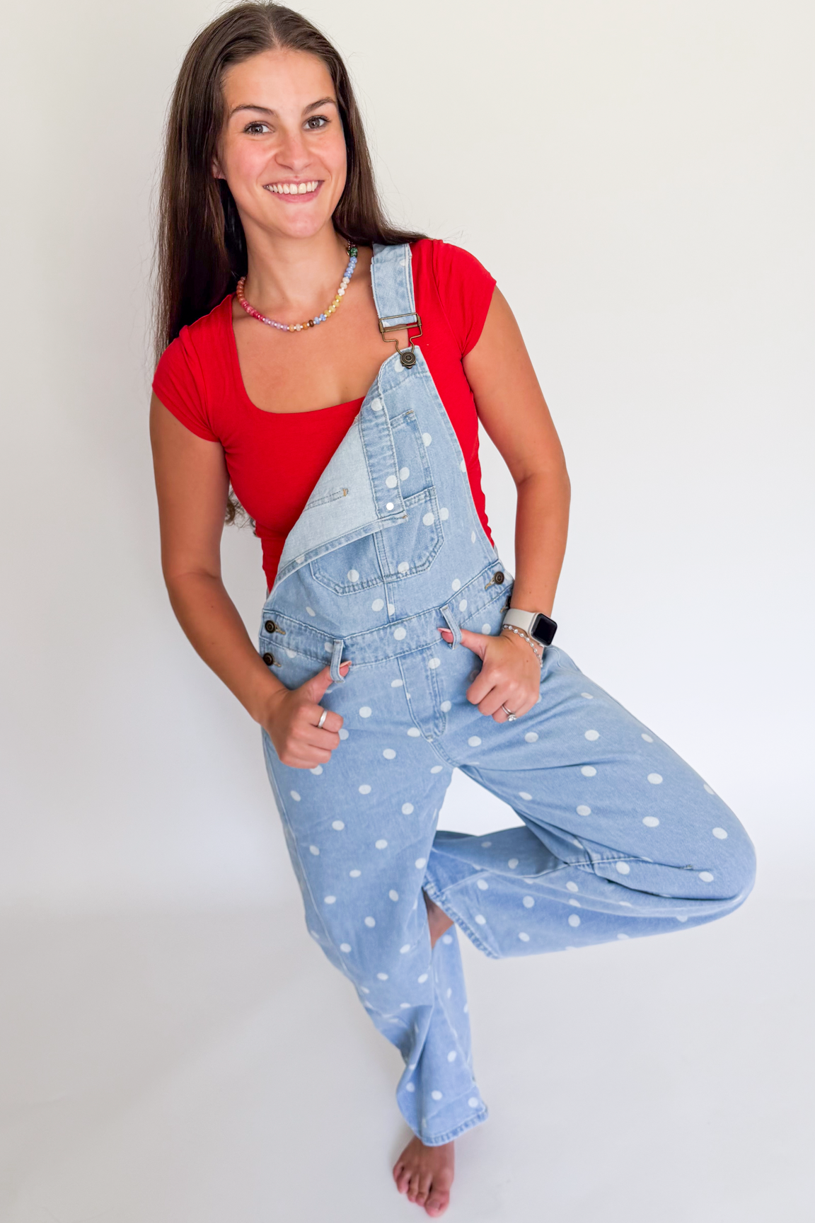 A woman posing in light blue denim overalls with white polka dots over a red top, standing barefoot against a plain white studio background.