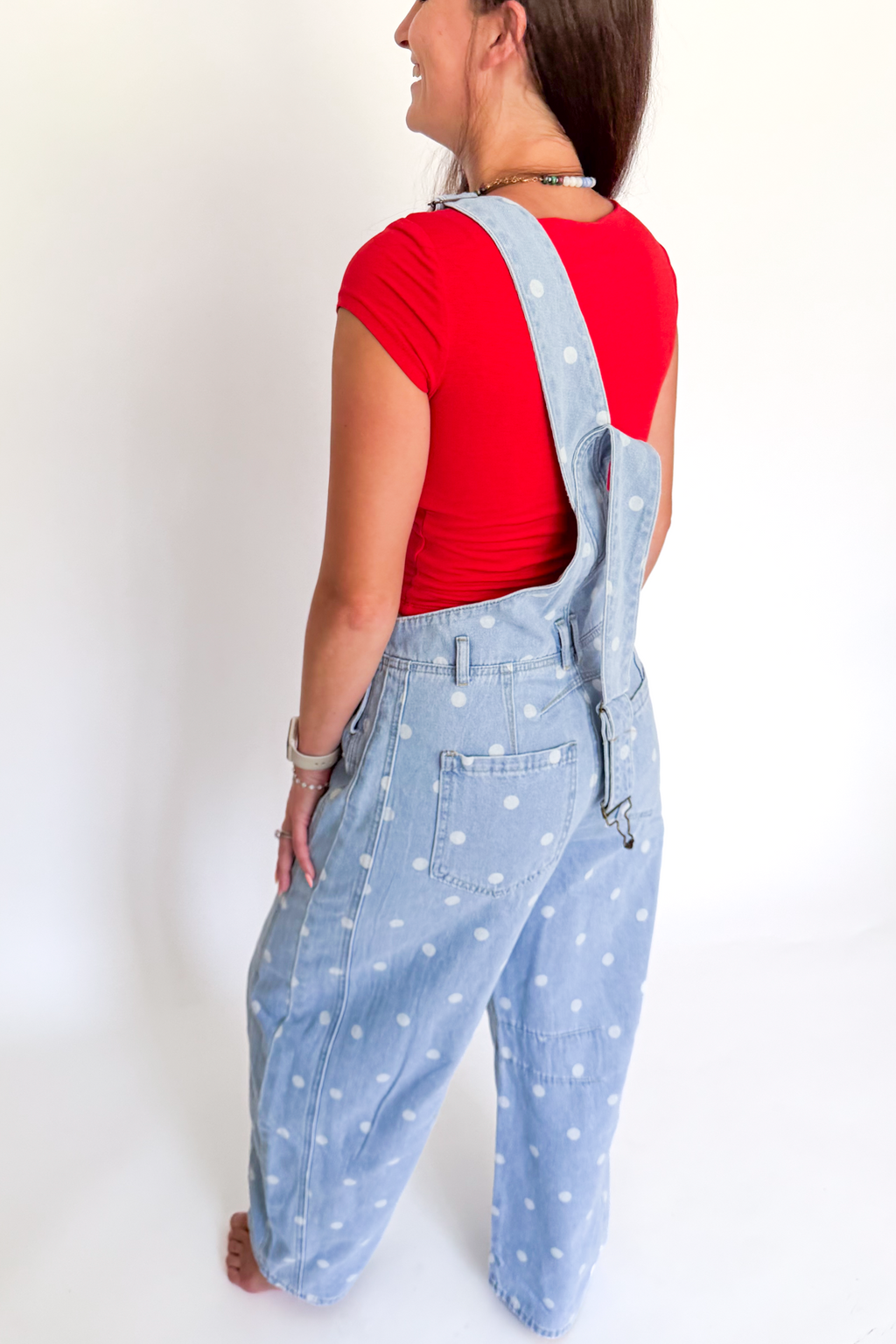 A woman wearing light blue denim overall with white polka dots, styled with a red t-shirt, shown from a side-back angle against a plain white background.