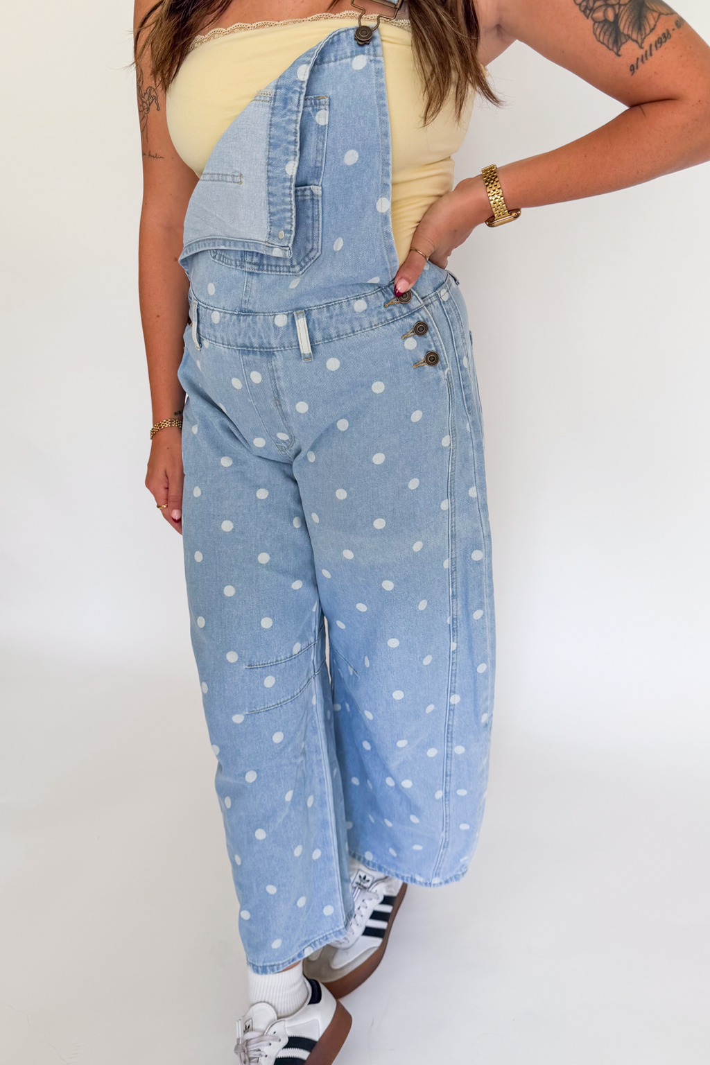 A person modeling light blue denim overalls with white polka dots, worn over a pale yellow top and white sneakers, against a plain white studio background.