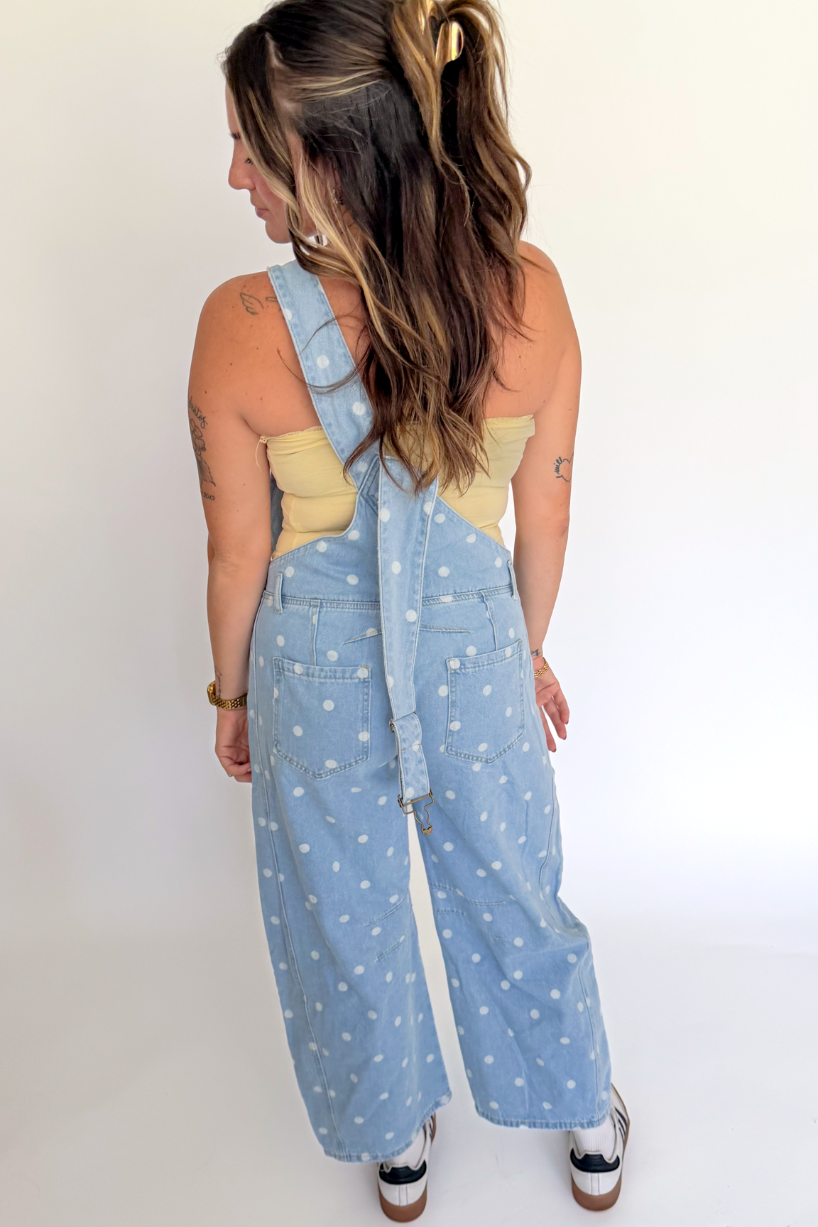 A woman shown from the back wearing a light blue denim overall with a white dalmatian spot pattern, styled with a beige bandeau top and black-and-white sneakers against a plain white studio background.