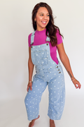 A woman modeling light blue denim overall with white polka dots over a bright pink t-shirt, standing against a plain white studio background.
