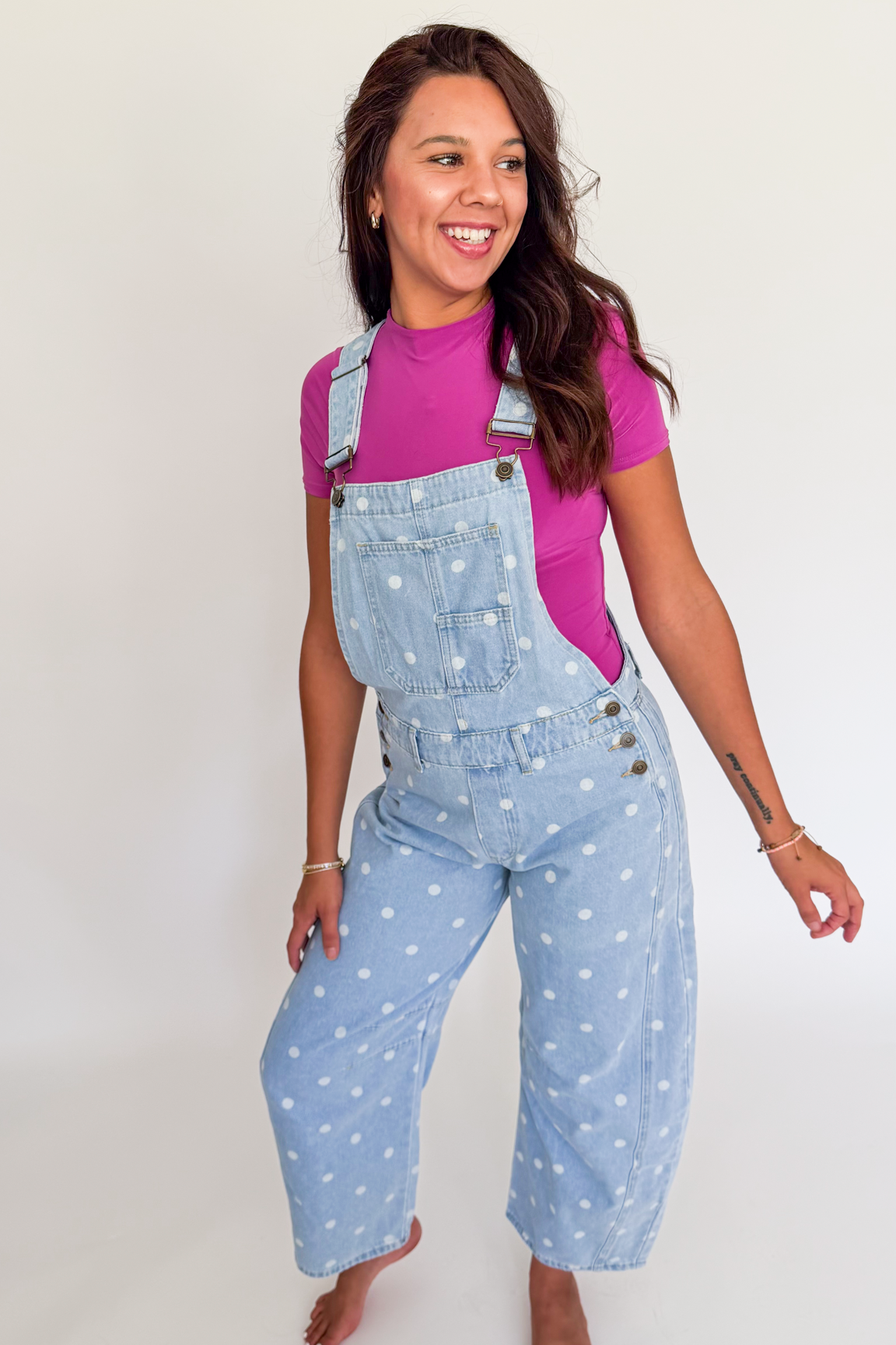 A woman modeling light blue denim overall with white polka dots over a bright pink t-shirt, standing against a plain white studio background.