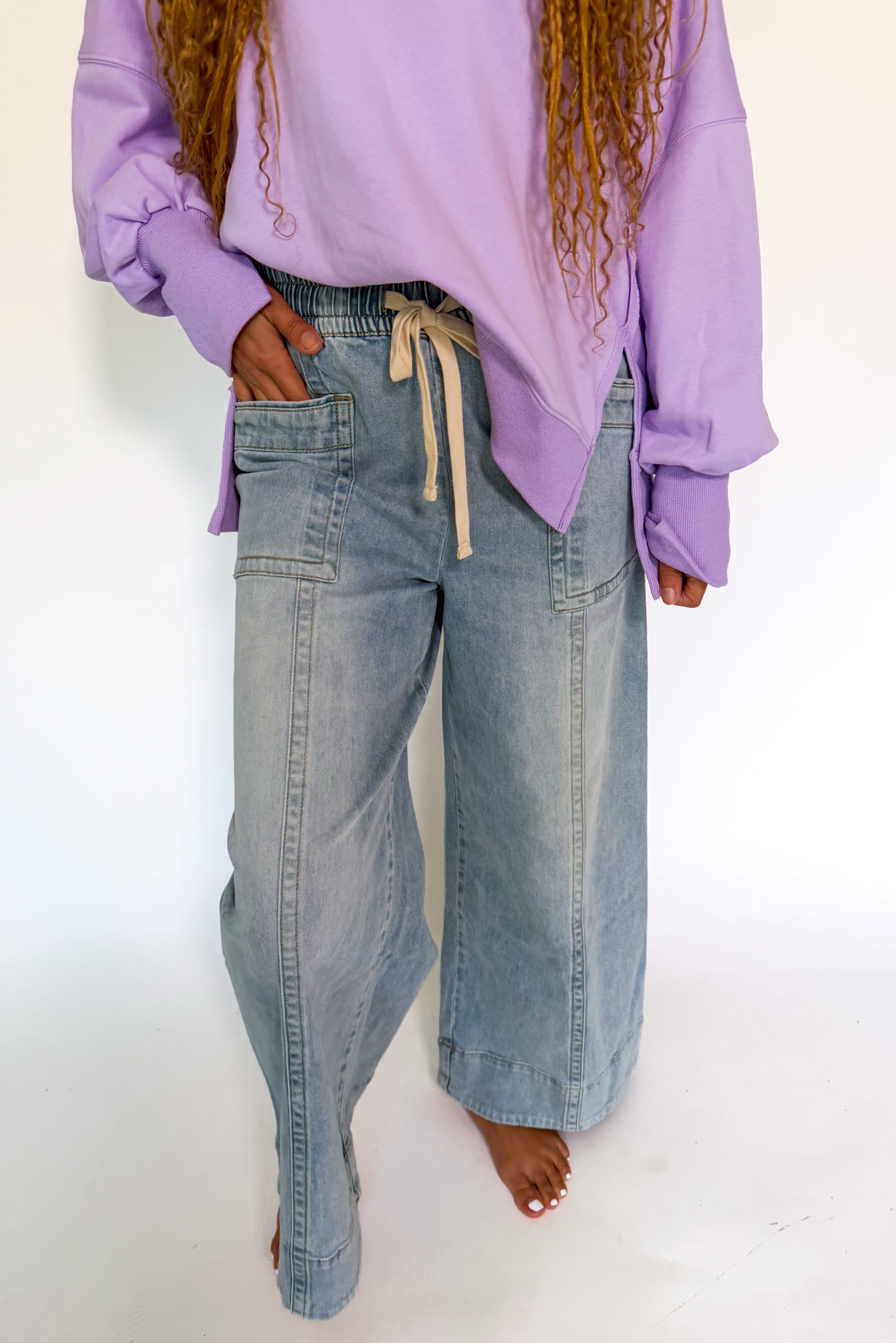 A person wearing light-wash wide-leg denim jeans with a cream drawstring waistband and a lavender sweatshirt, standing against a plain white studio background.