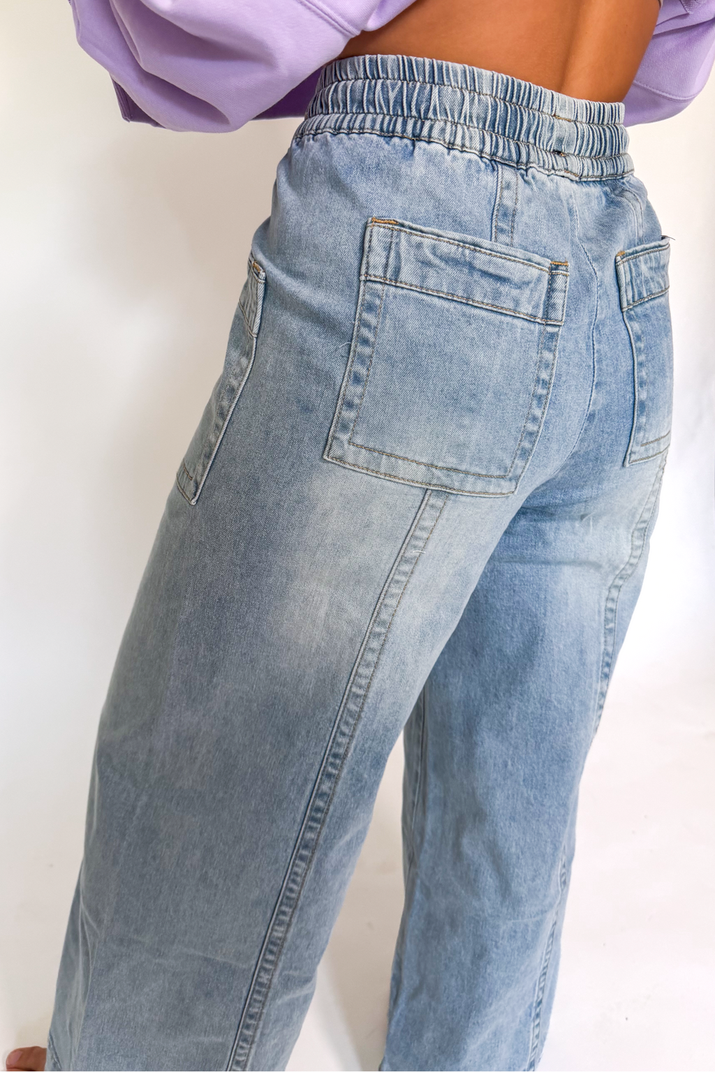 A person wearing light-wash blue denim jeans with an elastic waistband, shown from the back in a studio setting.