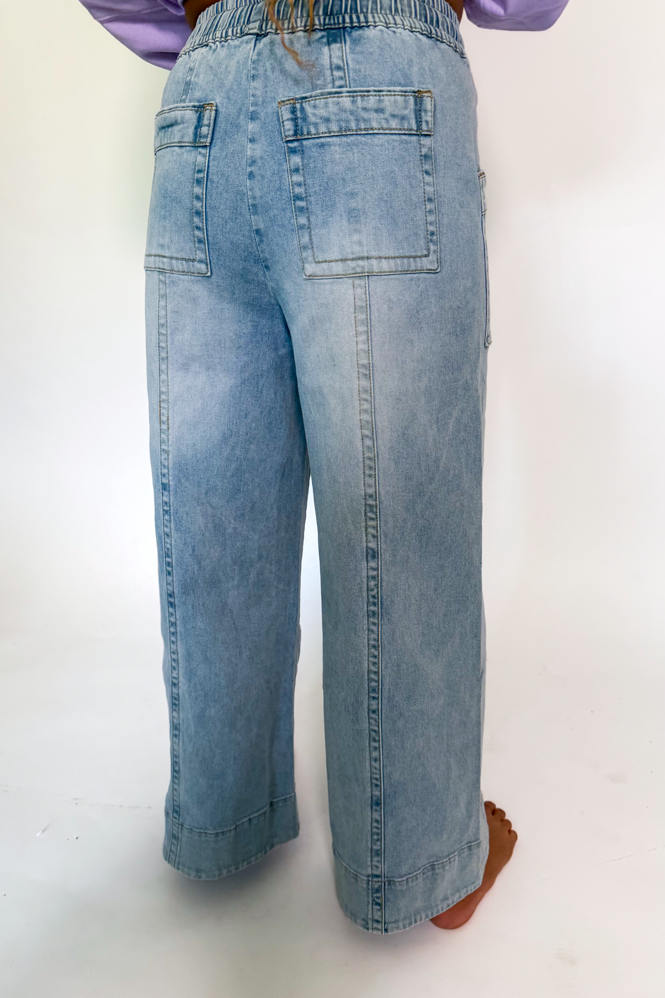 A person wearing light-wash blue denim wide-leg cropped pants, shown from the back against a plain white studio background.