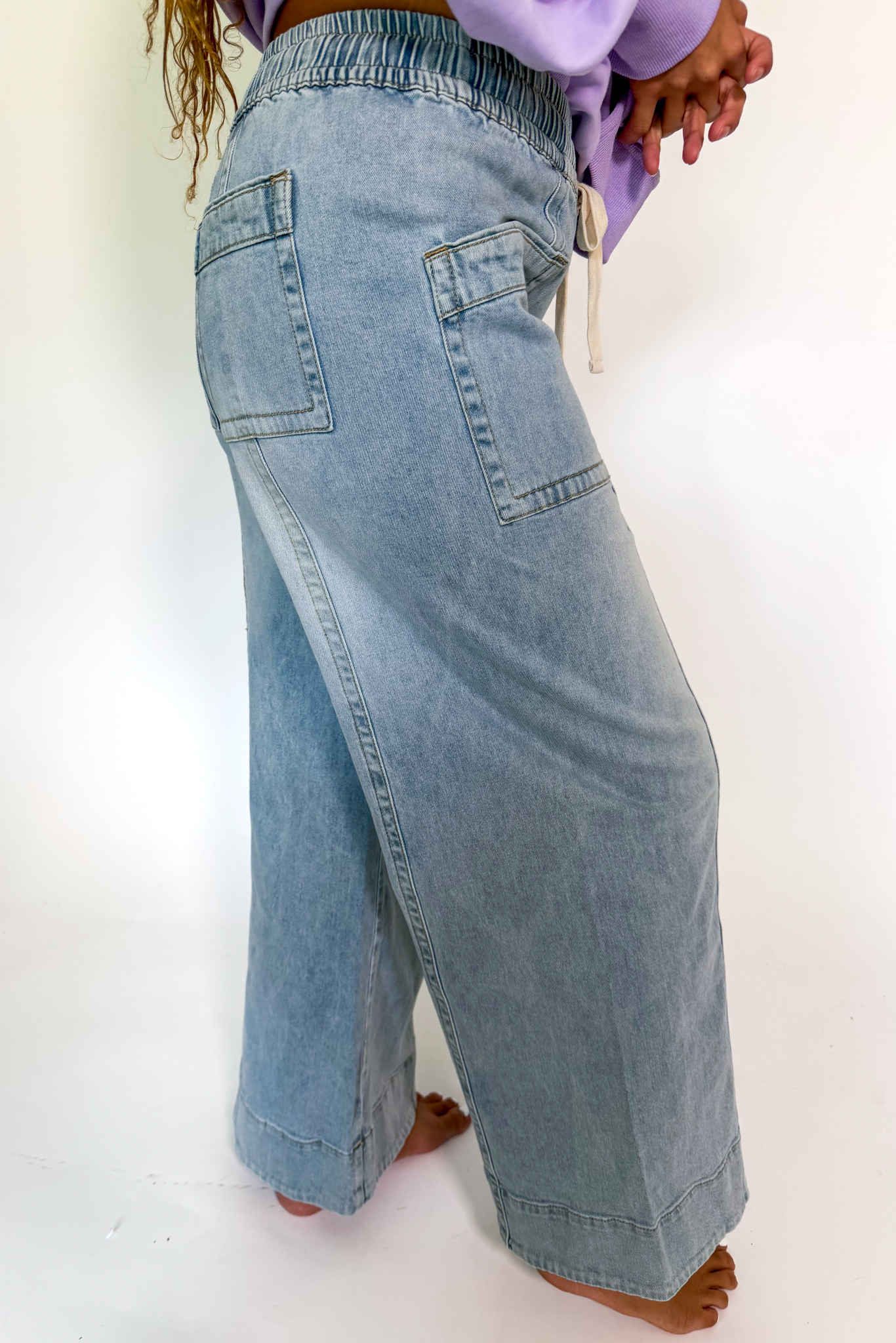 A person wearing wide-leg light-wash blue denim jeans with an elastic waistband and back patch pockets, shown from the back against a plain white studio background.