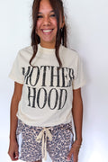 A smiling woman wearing a cream-colored graphic t-shirt with 'MOTHERHOOD' printed in distressed black letters, paired with leopard print shorts, against a plain white background.