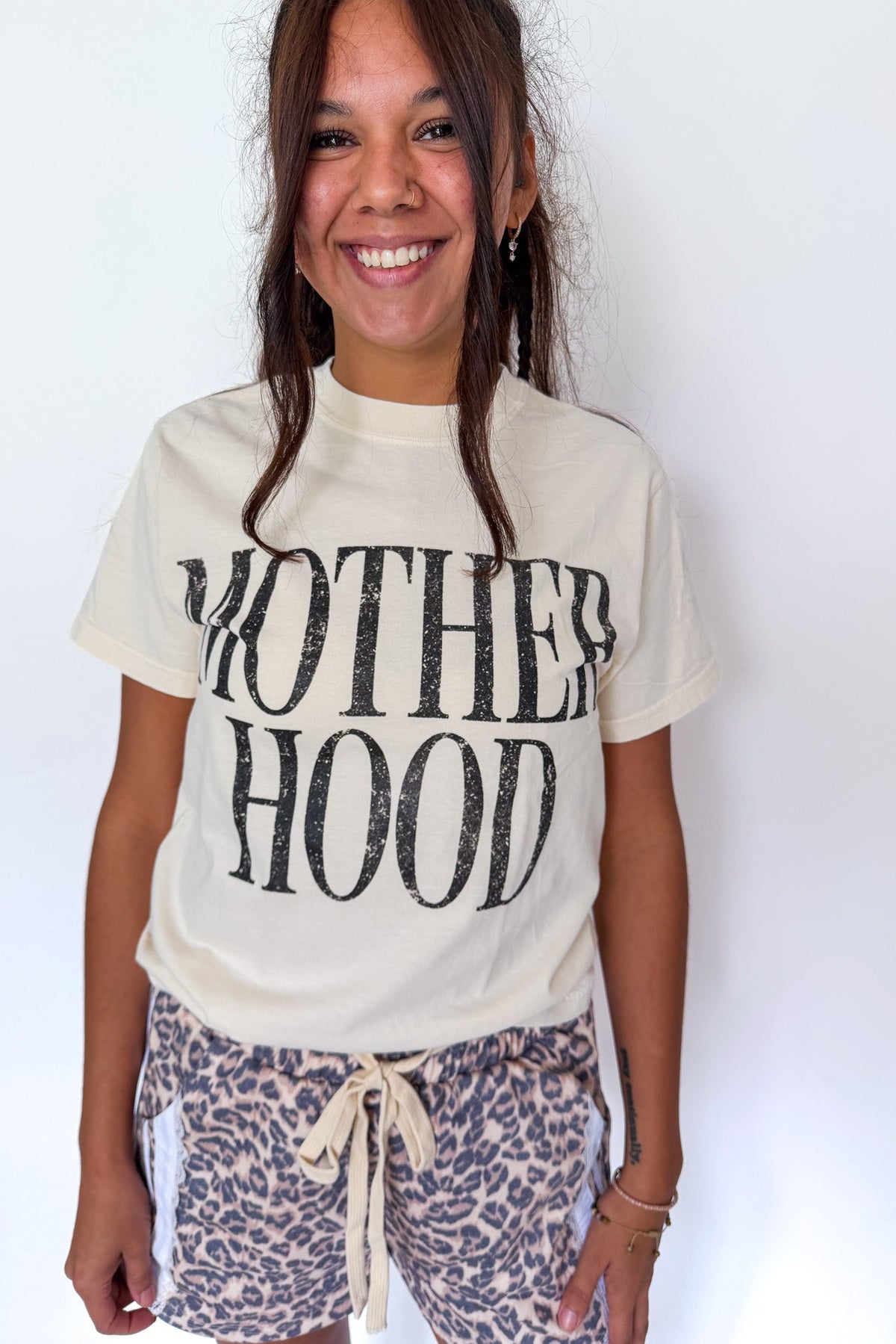 A smiling woman wearing a cream-colored graphic t-shirt with 'MOTHERHOOD' printed in distressed black letters, paired with leopard print shorts, against a plain white background.