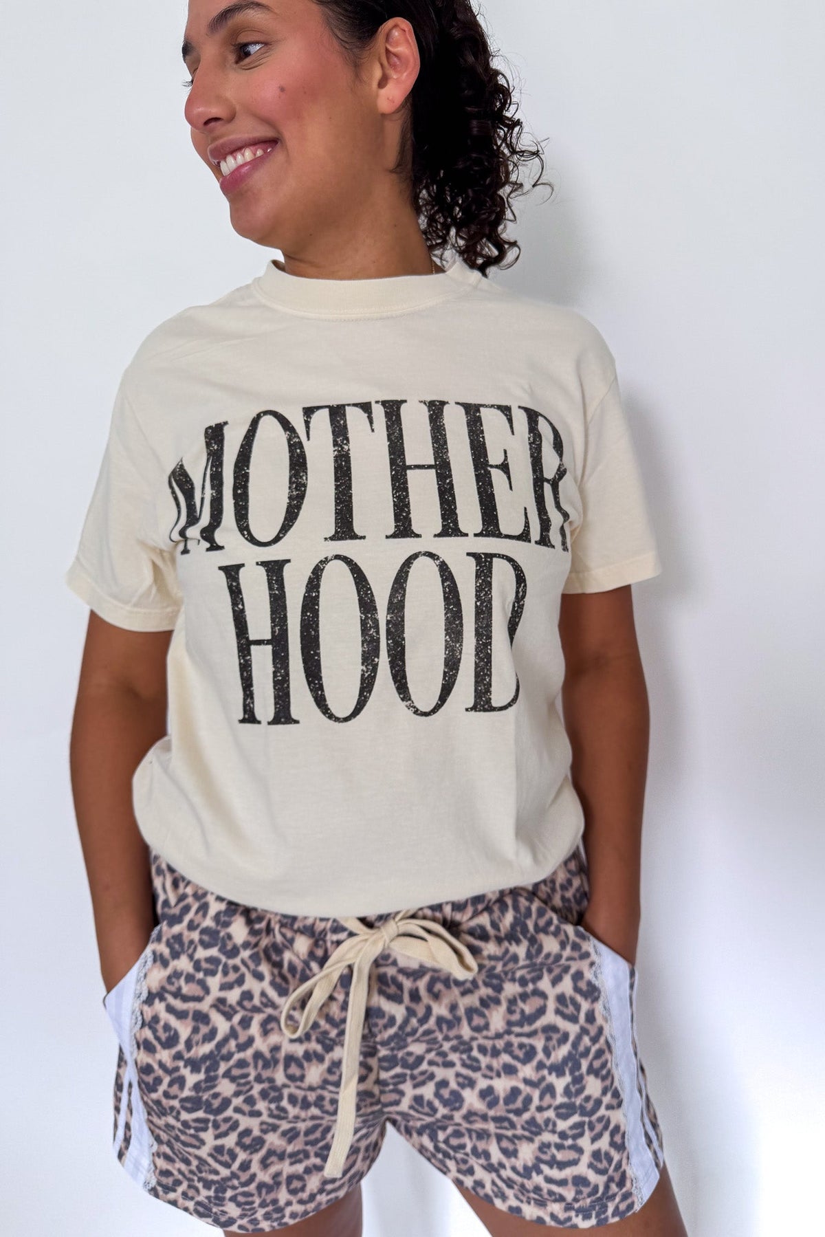 A woman wearing a cream-colored graphic t-shirt with 'MOTHERHOOD' printed in distressed black letters, paired with leopard print shorts, standing against a plain white wall.