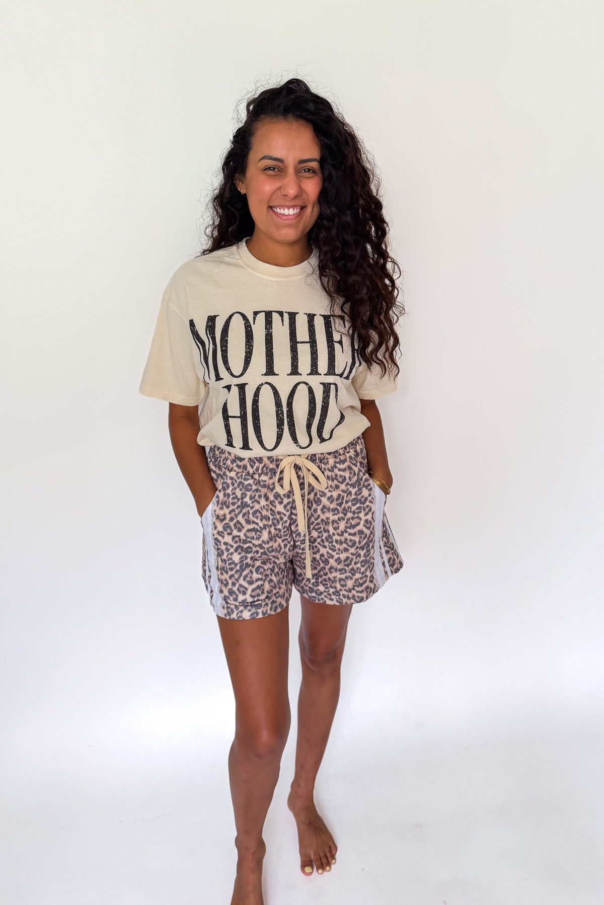 A woman modeling a beige graphic t-shirt that reads 'MOTHER HOOD' in black letters, paired with leopard print shorts featuring white side stripes, standing against a plain white background.