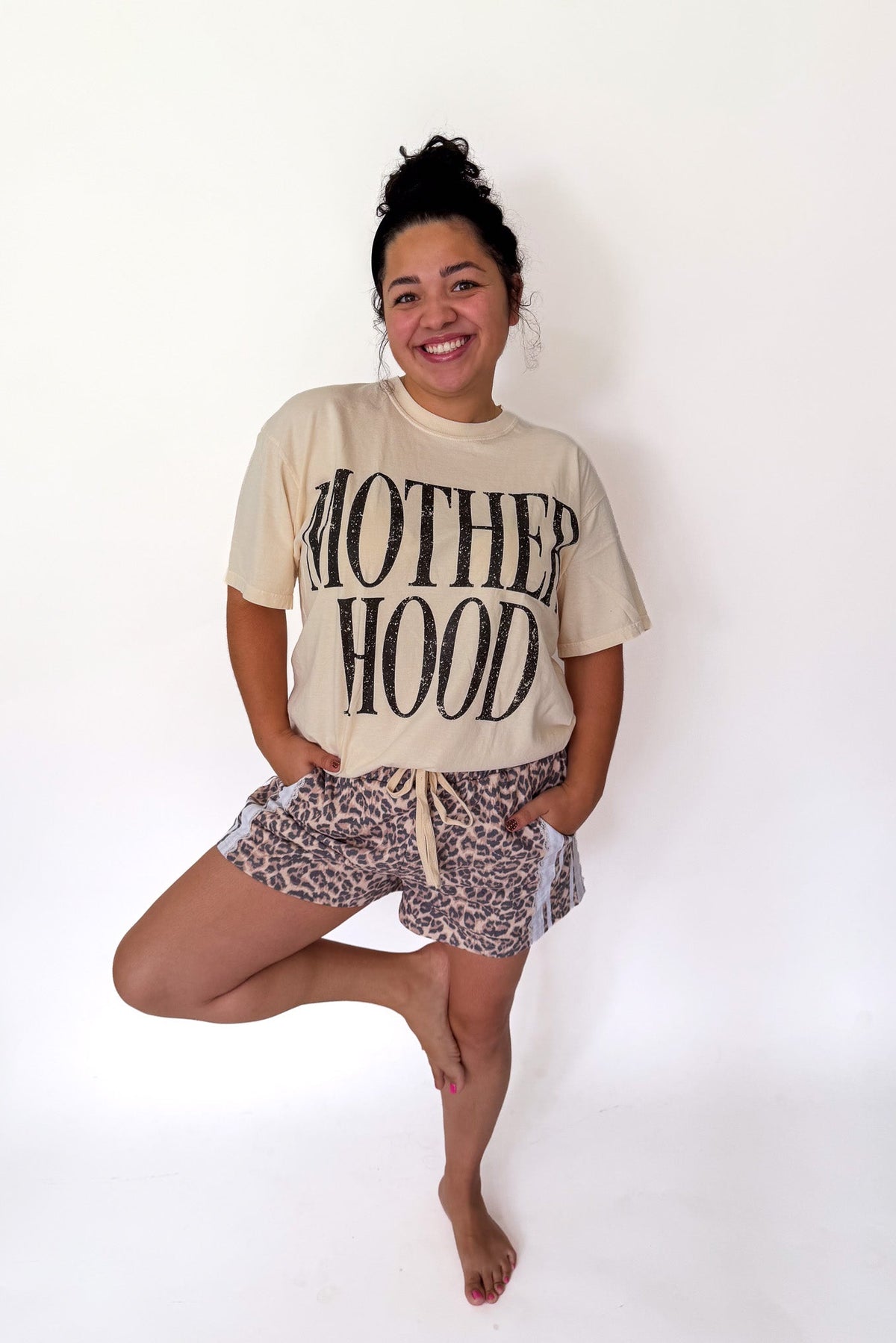 A woman posing in a cream graphic t-shirt with 'MOTHERHOOD' printed in black, paired with leopard print shorts, against a plain white background.