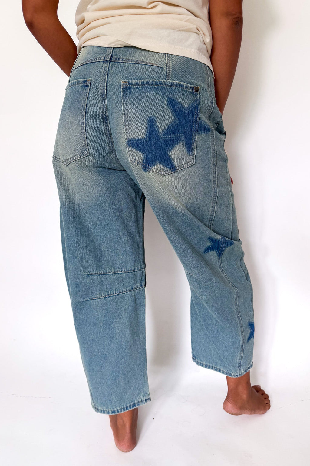 A person shown from the back wearing light-wash blue denim jeans with dark blue star-shaped patches on the back pockets, standing against a plain white background.