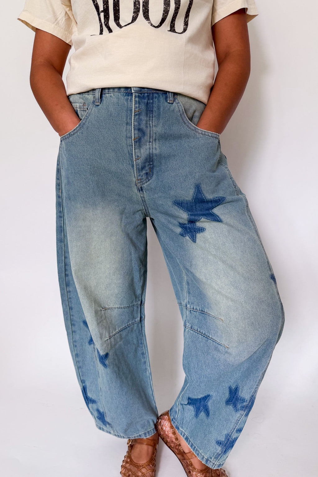 A person wearing light blue wide-leg denim jeans with dark blue star-shaped patch details on the front and lower legs, paired with a cream graphic t-shirt and brown sandals, against a plain white background.