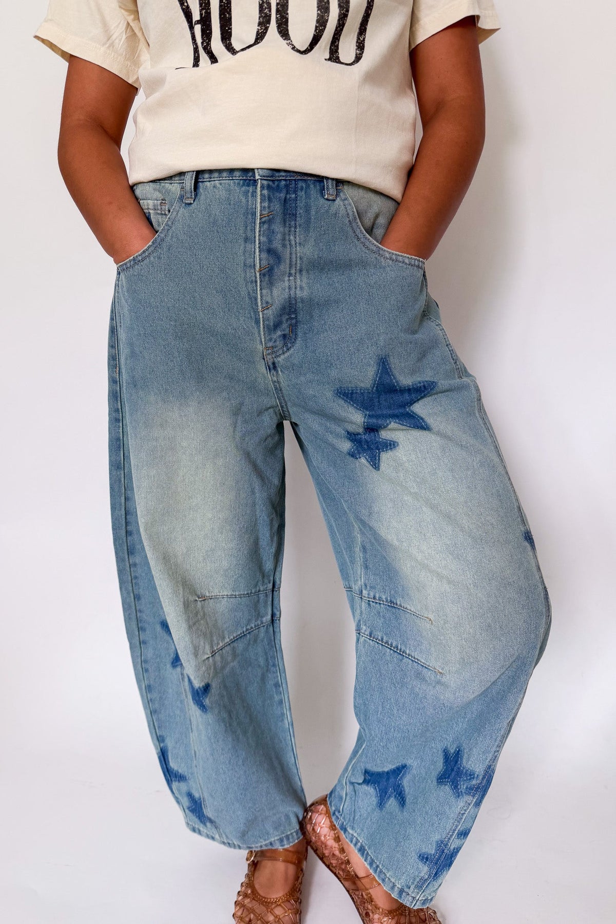 A person wearing light blue wide-leg denim jeans with dark blue star-shaped patch details on the front and lower legs, paired with a cream graphic t-shirt and brown sandals, against a plain white background.