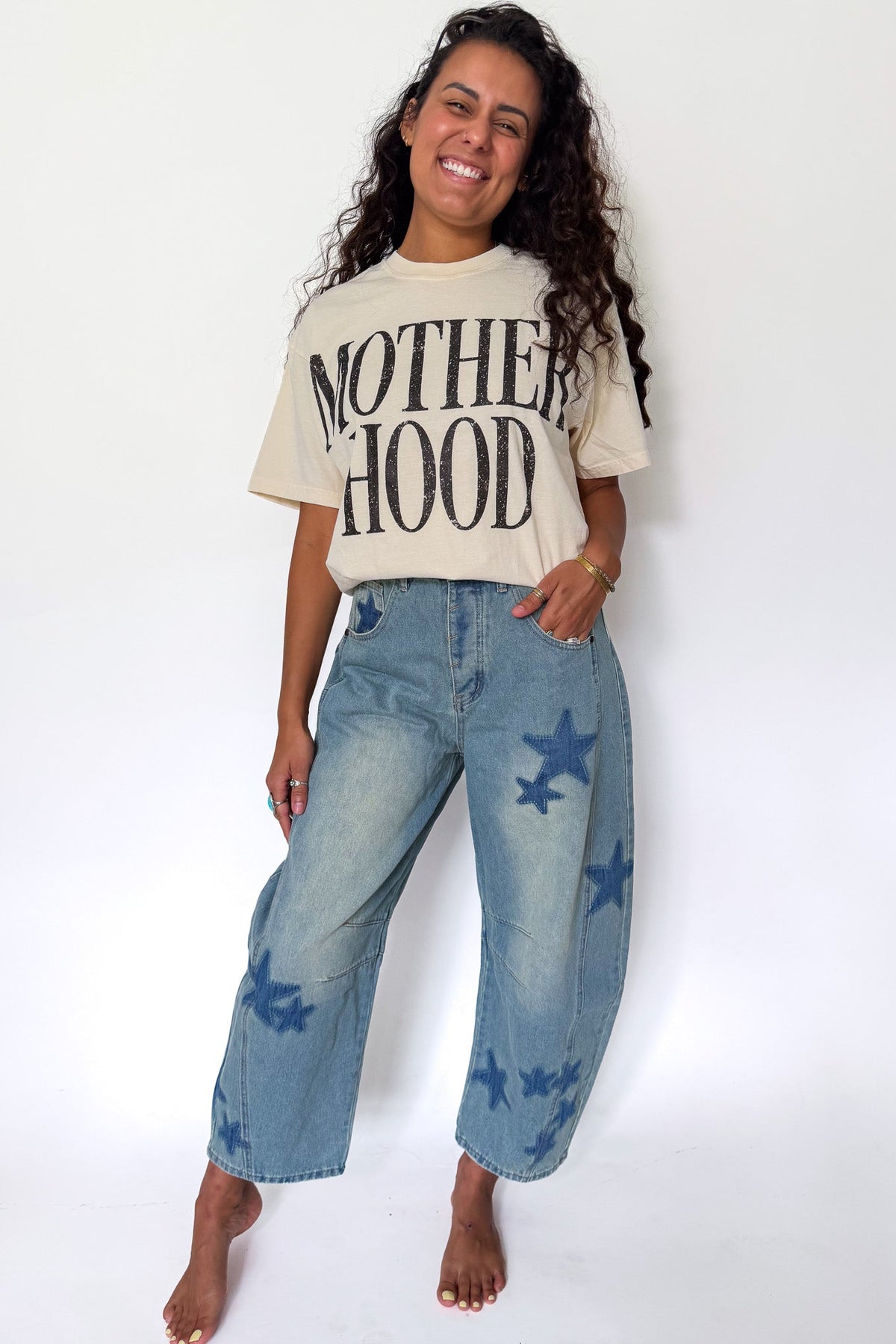 A woman stands barefoot against a plain white studio background, wearing a cream graphic T-shirt with the words 'MOTHERHOOD' in black and light-wash blue wide-leg jeans with dark blue star prints.