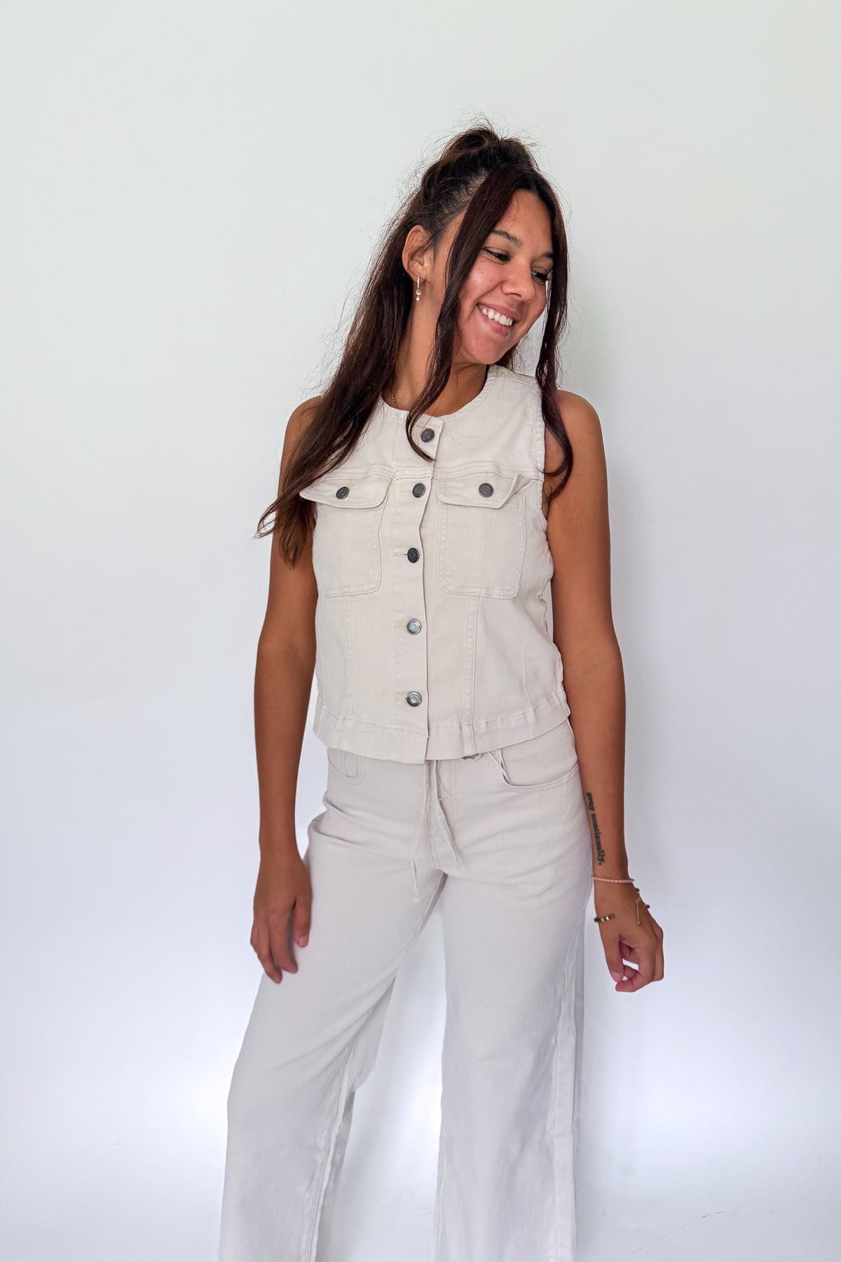 A woman modeling a sleeveless light beige denim vest with silver buttons and matching light beige wide-leg pants, standing against a plain white studio background.