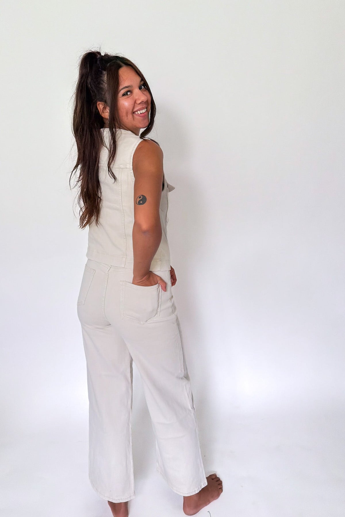 A woman with long dark hair in a ponytail, wearing a sleeveless cream-colored vest and matching wide-leg pants, posing against a plain white studio background.