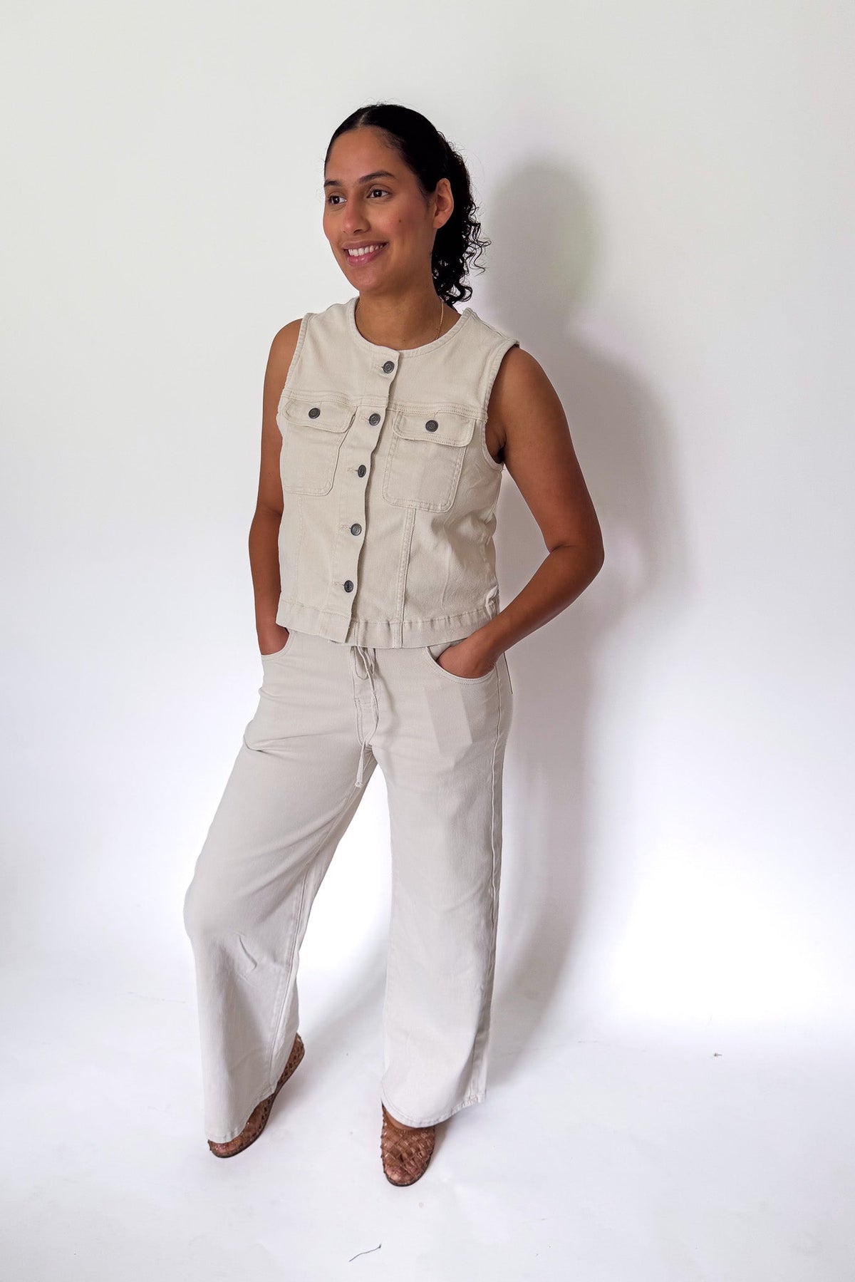 A woman modeling a light beige sleeveless button-front vest and matching wide-leg pants against a plain white studio background.