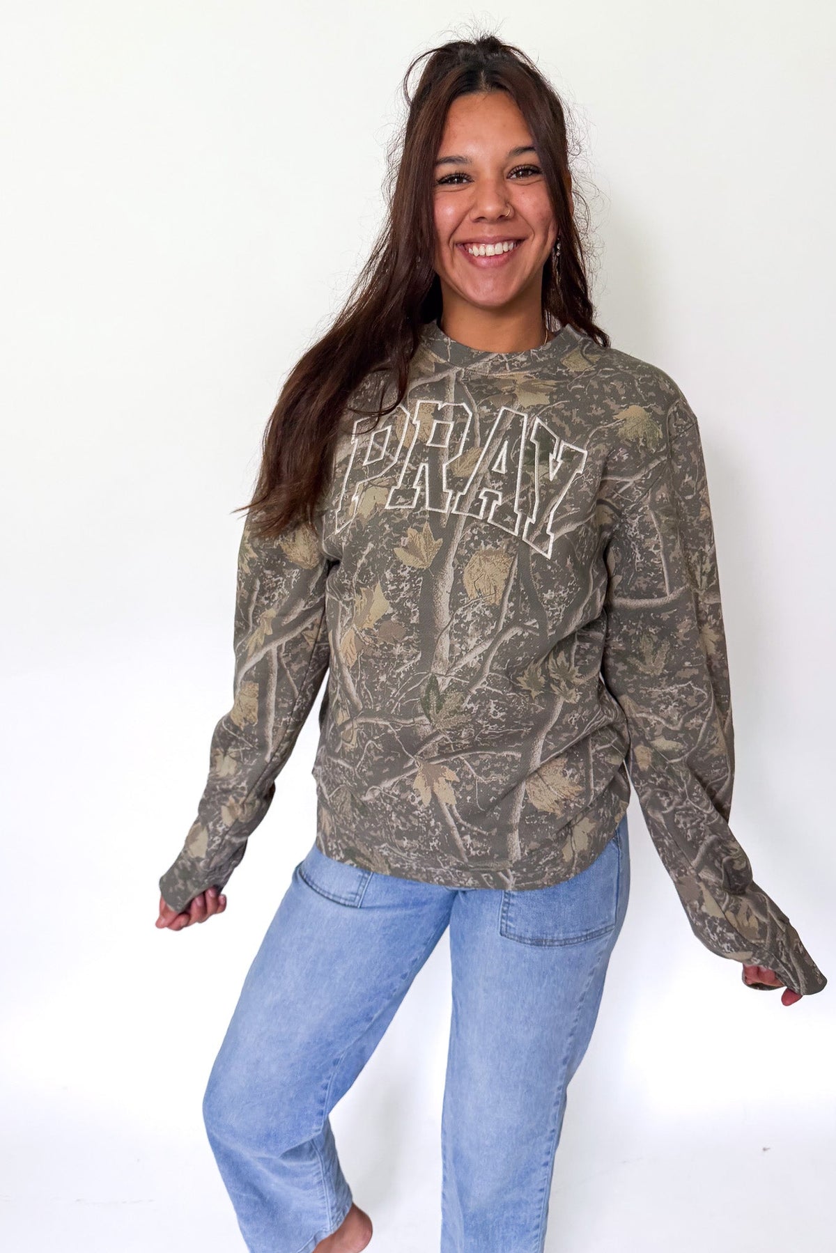 A woman modeling a camouflage long-sleeve hoodie with 'ARMY' printed in white arched letters across the chest, paired with light blue jeans, against a plain white background.