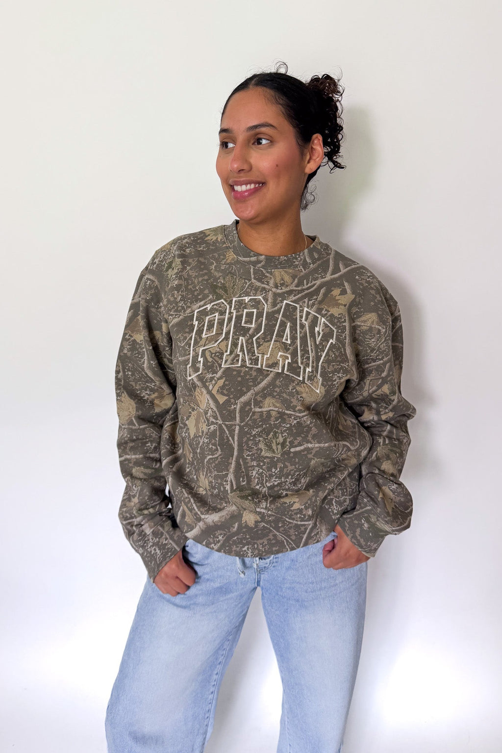 A woman wearing a brown and gray camouflage crewneck sweatshirt with 'PRAY' printed in white arched letters on the chest, paired with light blue jeans, standing against a plain white wall.