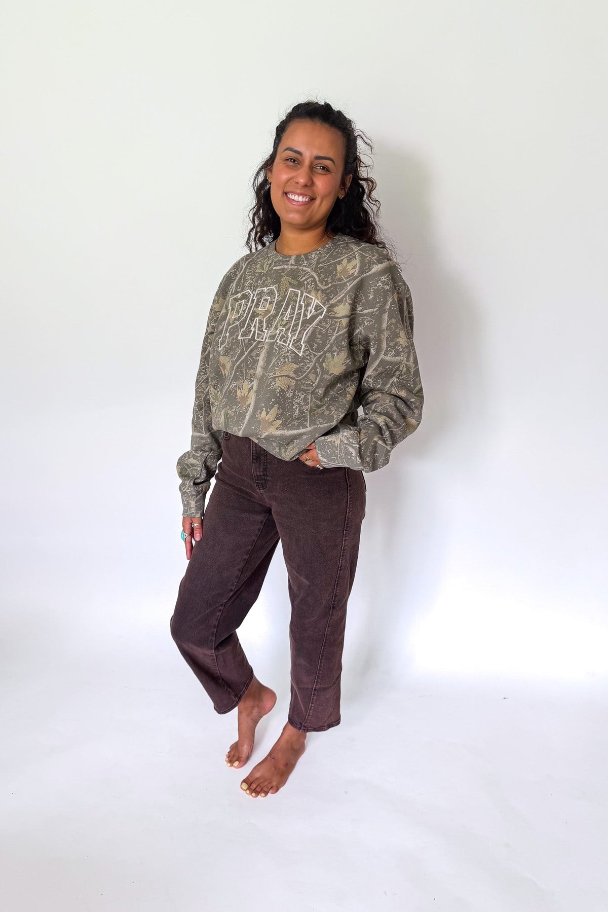 A woman stands barefoot against a white studio background wearing a faded olive-green graphic sweatshirt and dark brown cropped pants.