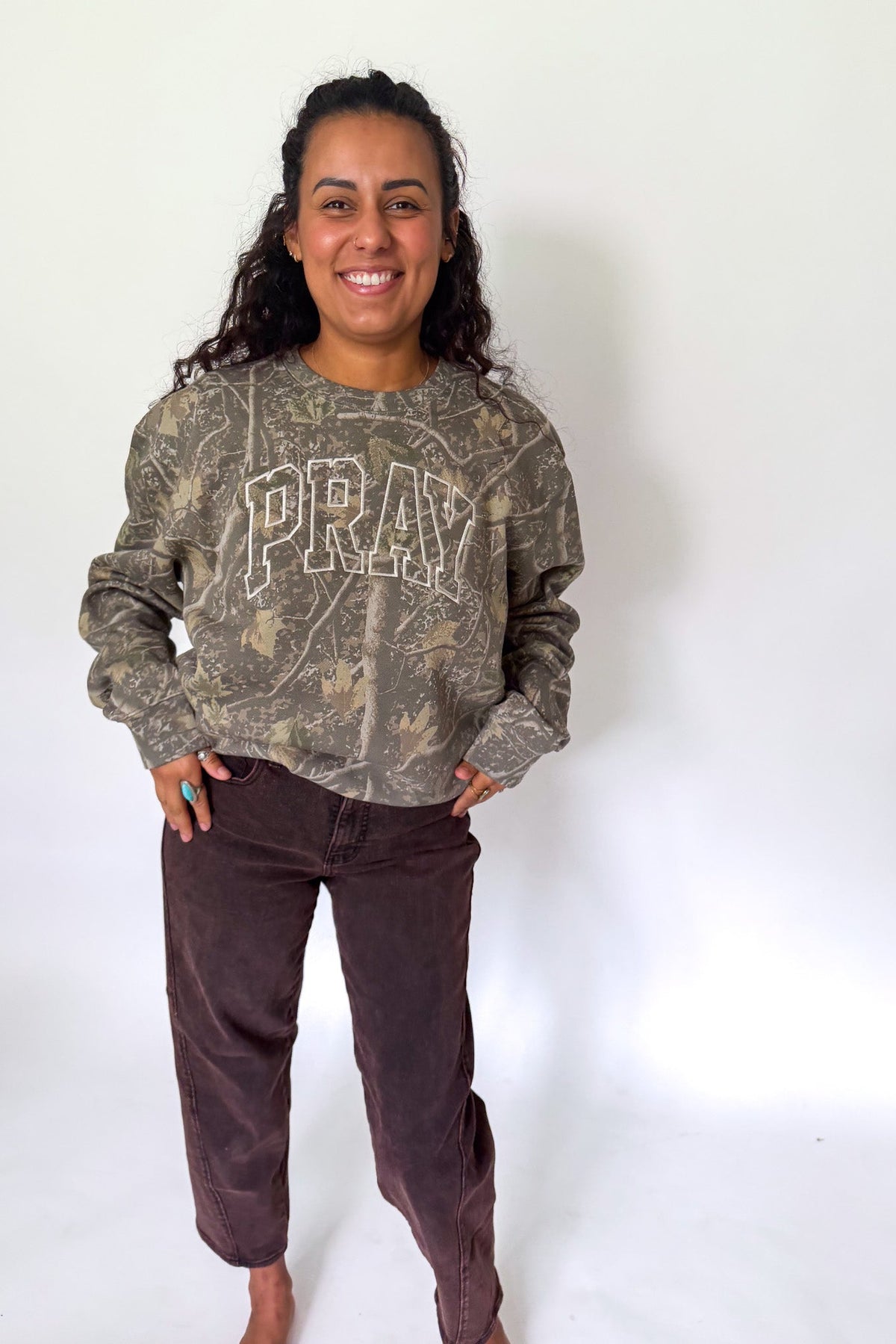 A woman standing against a white studio background wearing a long-sleeve crewneck sweatshirt with a distressed camouflage pattern and the word 'PRAY' printed across the chest, paired with dark brown pants.